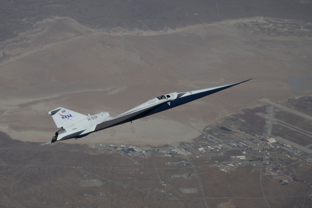 NASA’s X-59 quiet supersonic research aircraft flies high above the Mojave Desert. The aircraft is shown from a side profile with its long nose, swept wings and white fuselage stand out against the background.