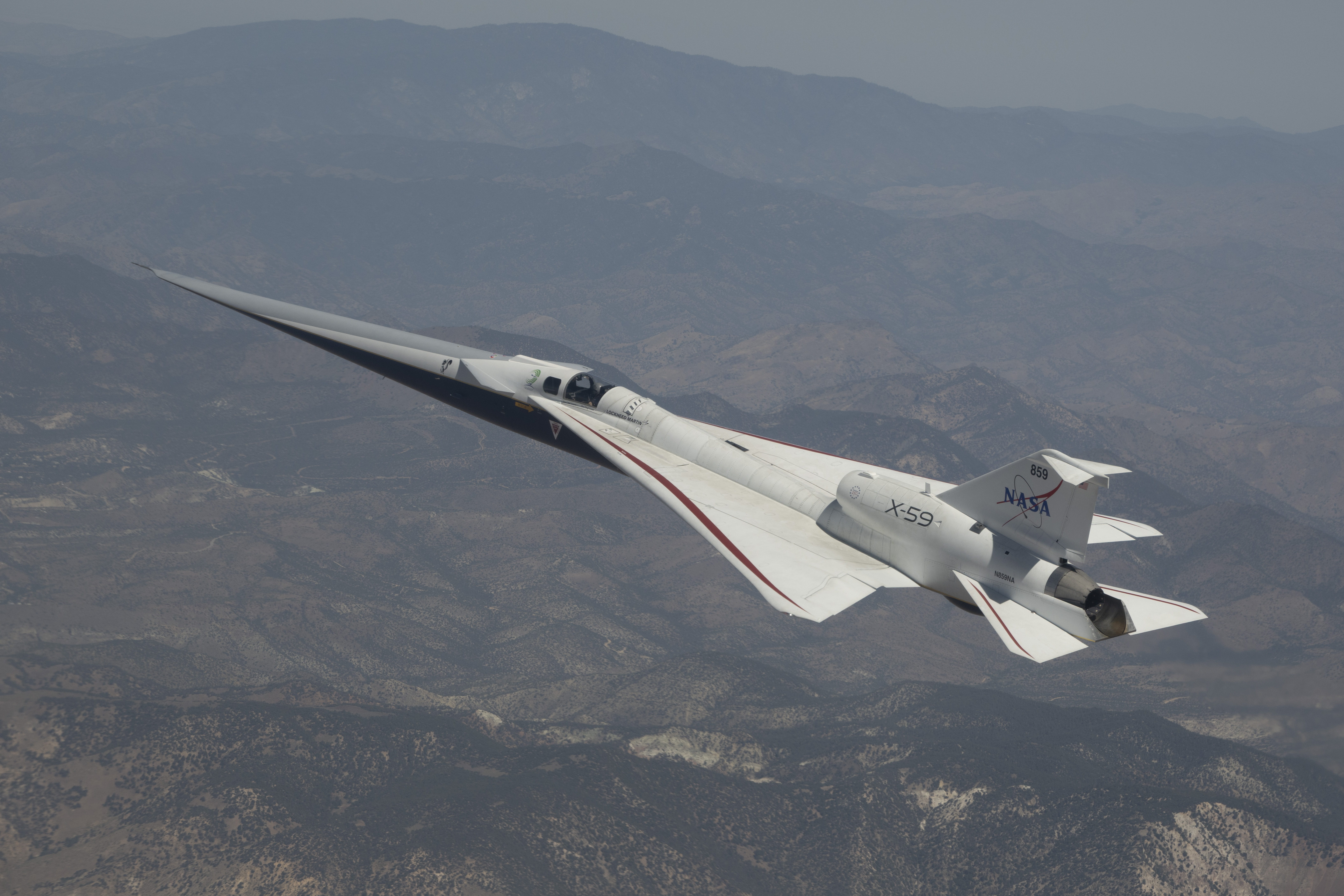 NASA’s X-59 quiet supersonic research aircraft is shown flying above mountain ridges in California during a test flight on May 12, 2026. The aircraft is viewed from above, showing its elongated nose, swept wings, and white fuselage against layered mountains and valleys below.
