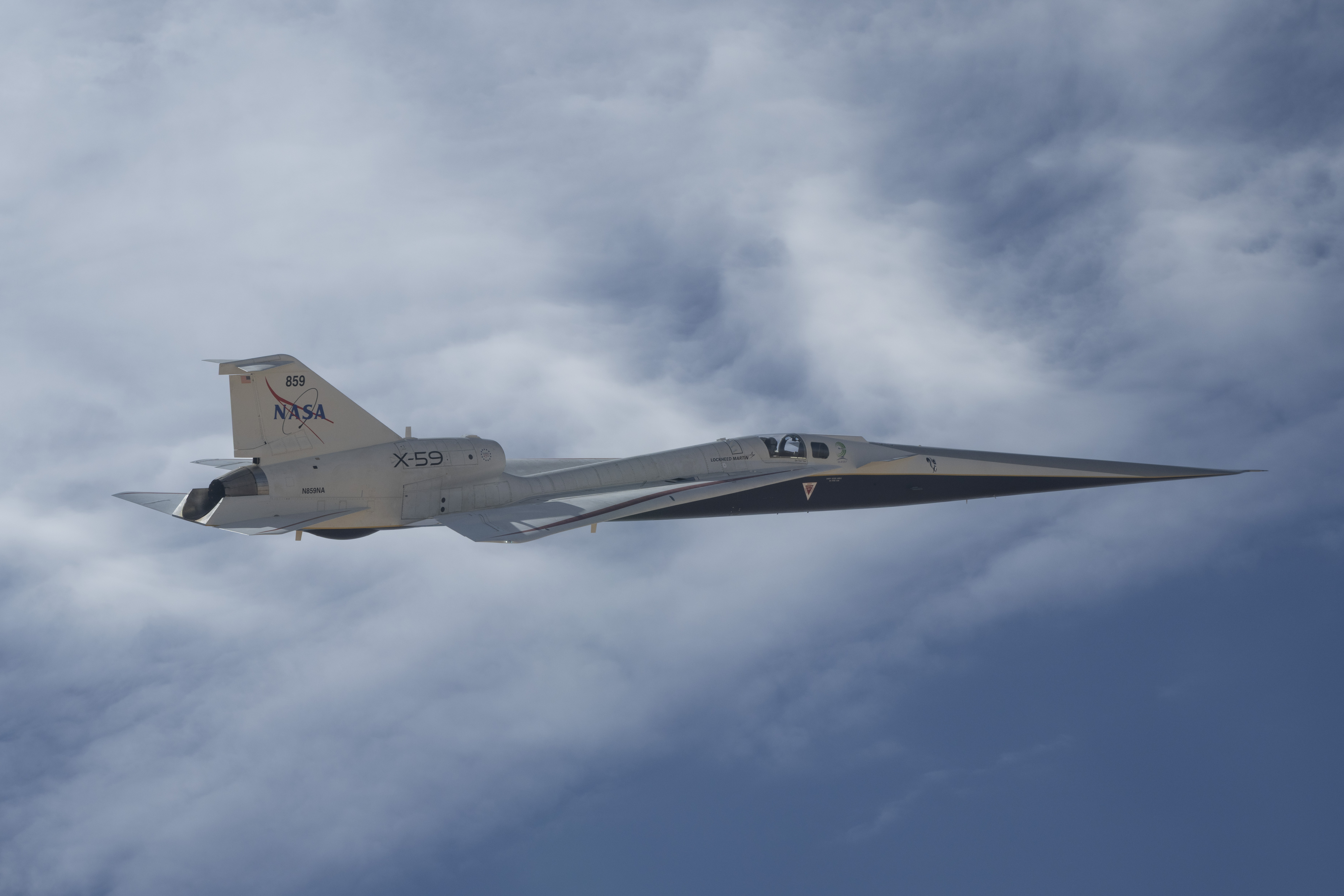 NASA’s X-59 quiet supersonic research aircraft is shown flying alongside clouds during a test flight on May 12, 2026. Viewed from the side, the aircraft’s length, swept wings and white fuselage stand out against patches of blue sky and clouds.