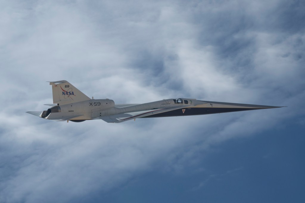 NASA’s X-59 quiet supersonic research aircraft is shown flying alongside clouds during a test flight on May 12, 2026. Viewed from the side, the aircraft’s length, swept wings and white fuselage stand out against patches of blue sky and clouds.