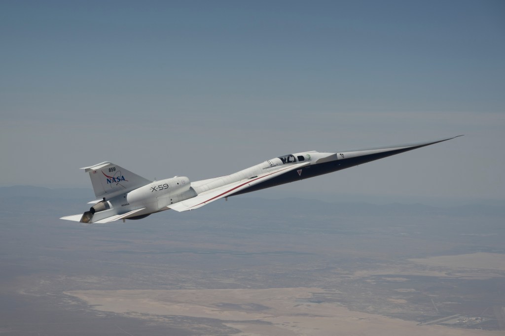 NASA’s X-59 quiet supersonic research aircraft is shown flying above mountain ridges in California during a test flight on May 12, 2026. The aircraft is viewed from above, showing its elongated nose, swept wings, and white fuselage against layered mountains and valleys below.
