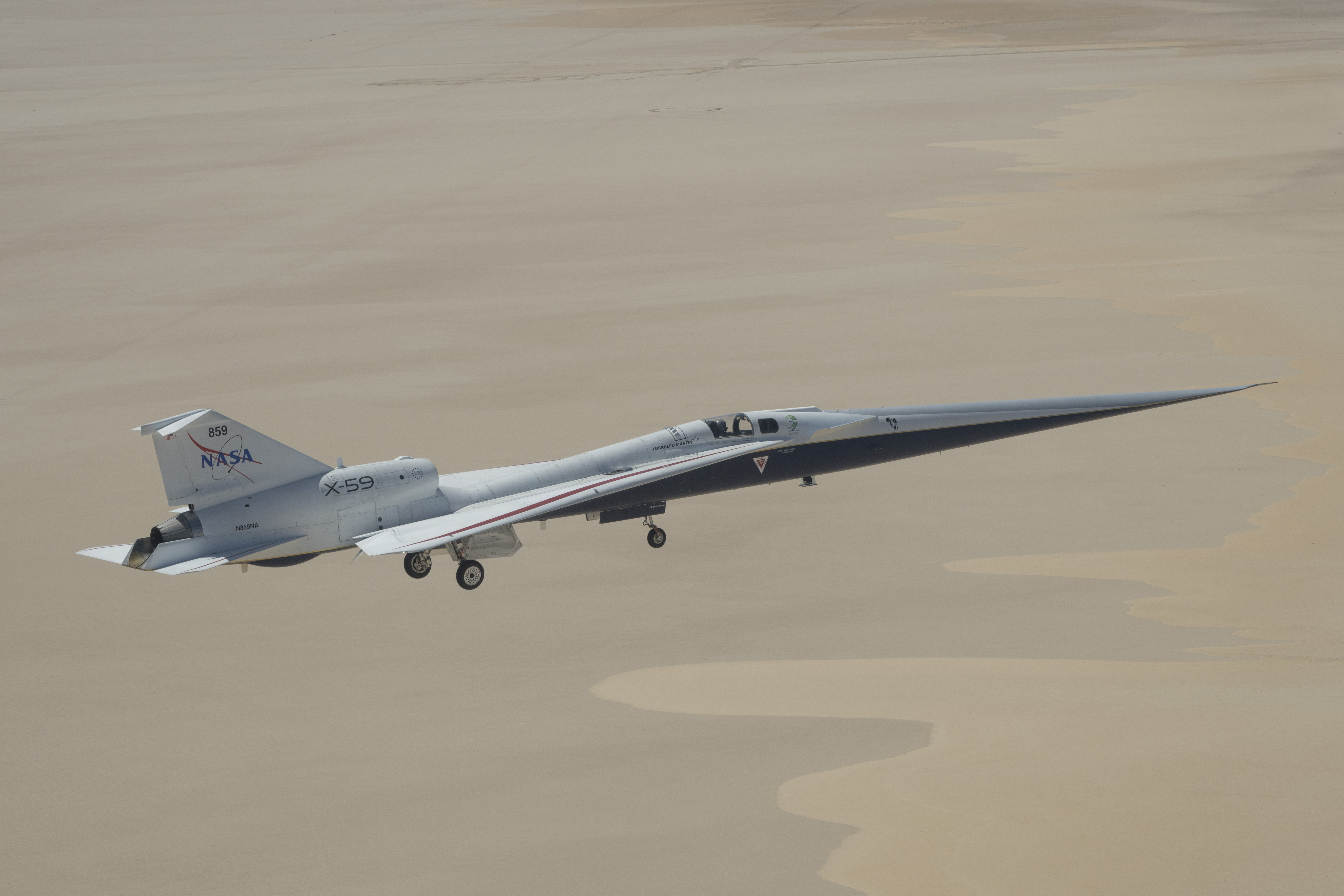 NASA’s X-59 quiet supersonic research aircraft flying above Rogers Dry Lake in California with its landing gear extended during a test flight on May 12, 2026. The aircraft’s long nose and white fuselage are visible against the light tan desert surface below.