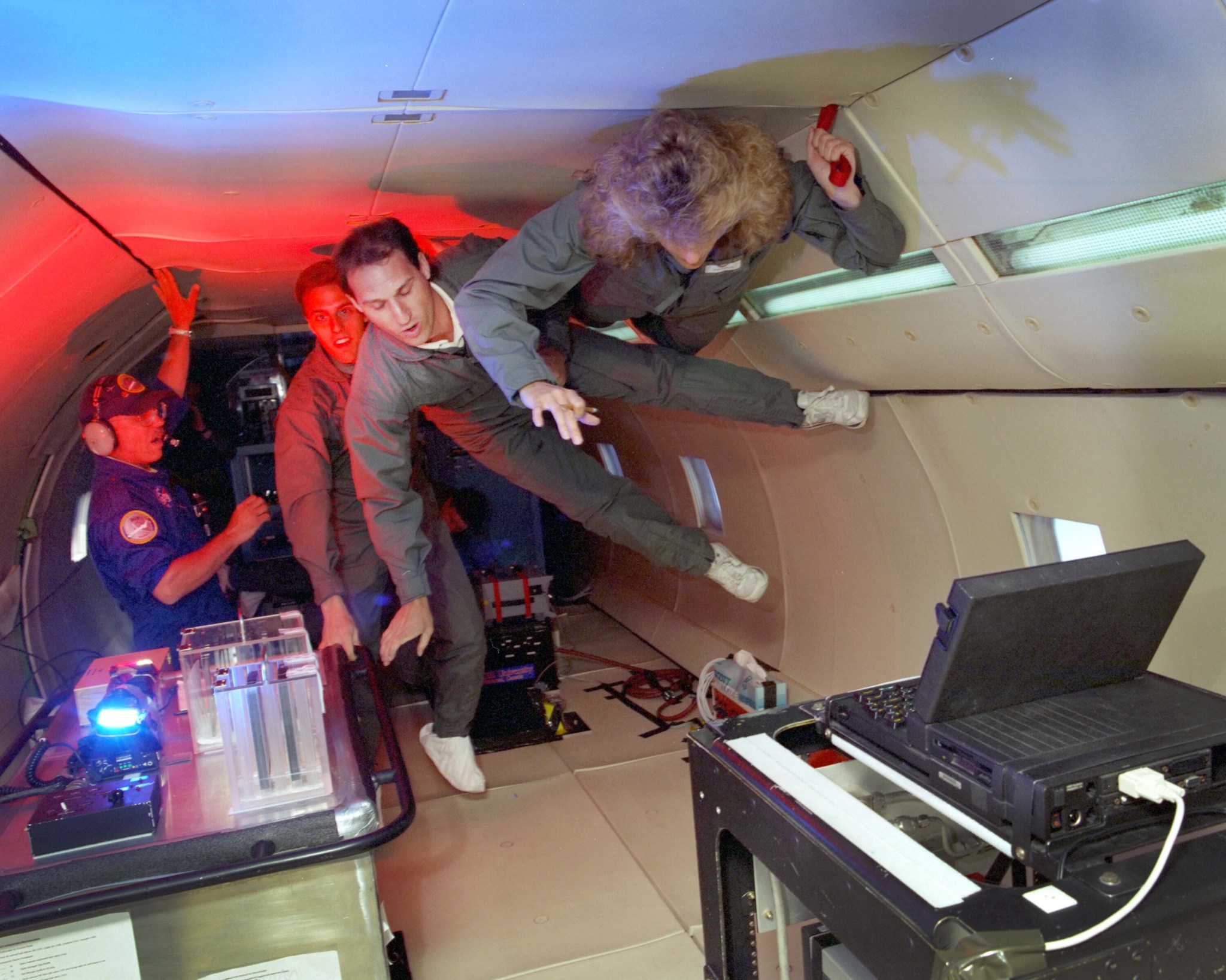 Inside the bed of an aircraft with no seating, three people in gray flight suits hang onto the sides of the plane to gain balance in the microgravity atmosphere. To the left, a flight research staff member assists. Researchers make their way to two small tables that contain test hardware.