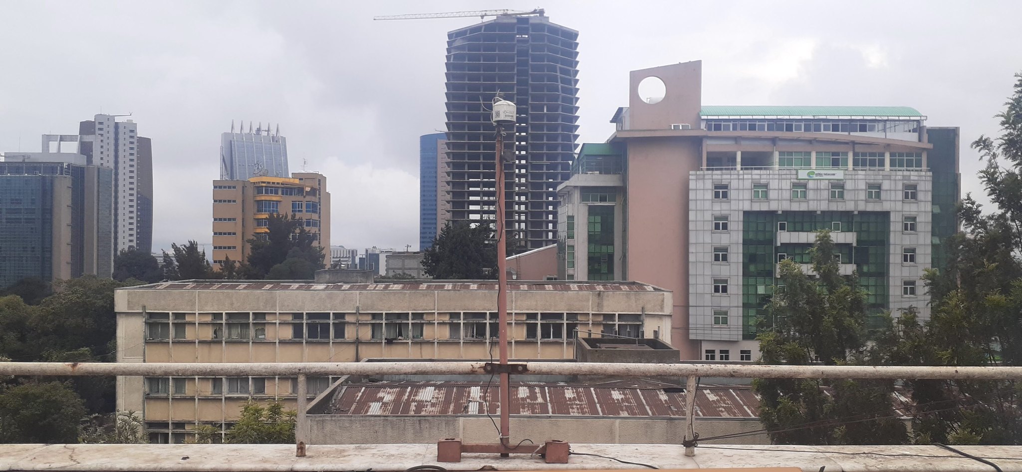 An air quality monitoring sensor mounted on a rooftop stands in front of a dense urban skyline, capturing atmospheric data. The sensor is semi-cylindrical white object several inches across, mounted on a tall rust-colored pole.