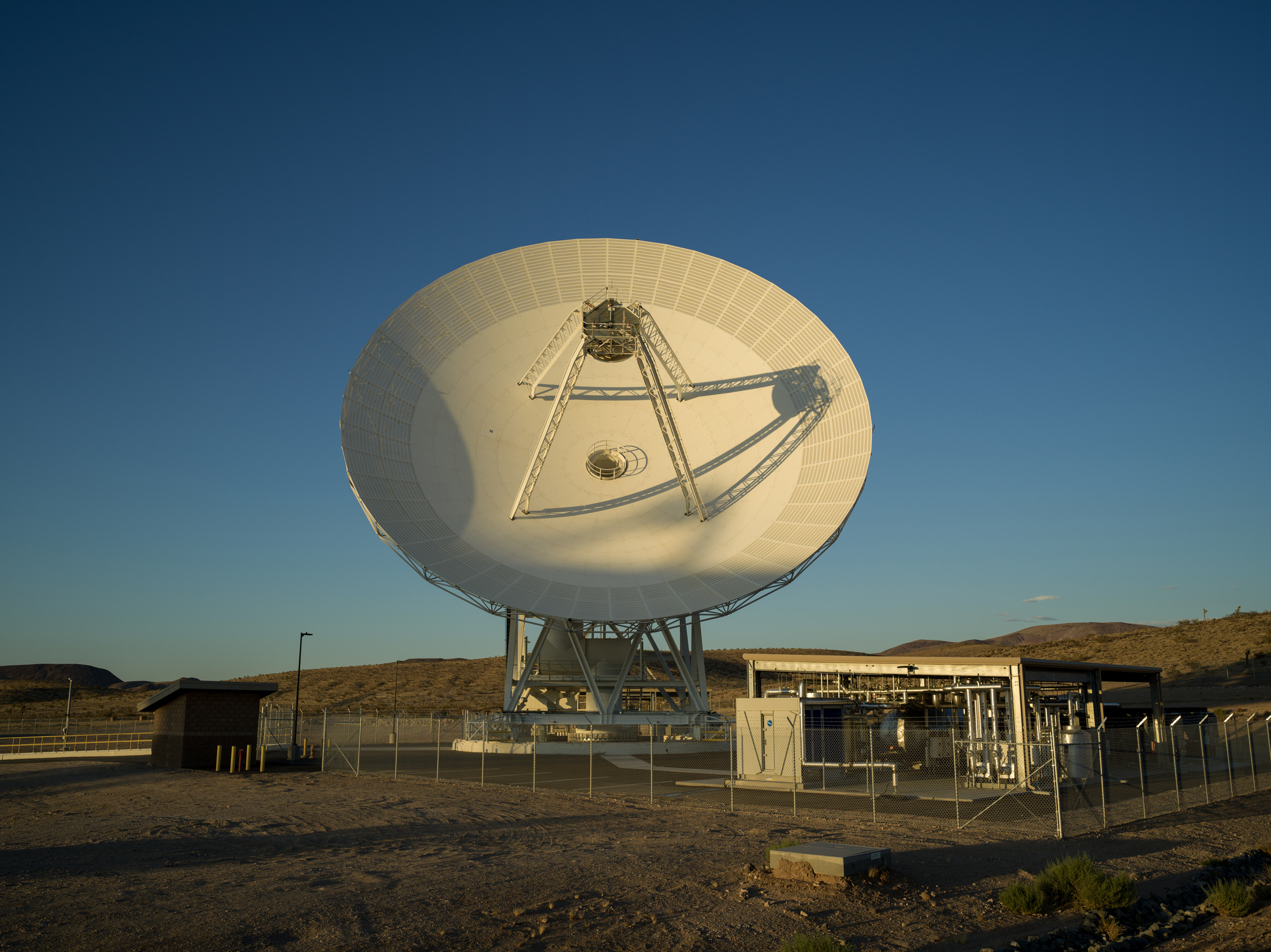 A massive white satellite dish antenna stands on a desert plain under a clear blue sky, bathed in warm sunlight alongside small facility structures.