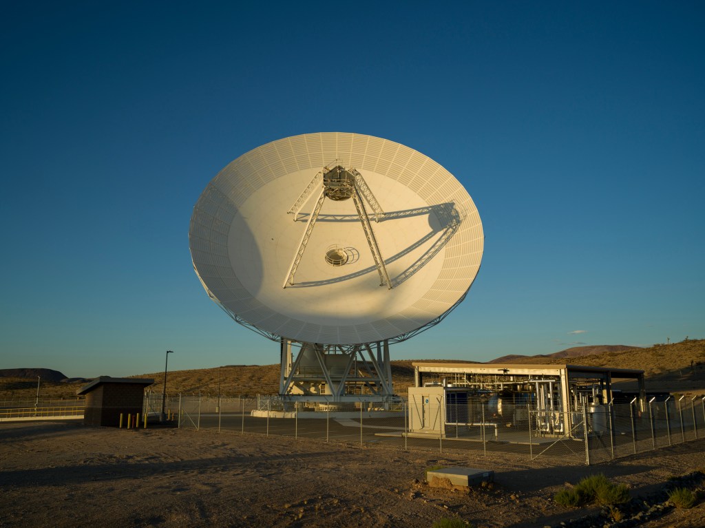 A massive white satellite dish antenna stands on a desert plain under a clear blue sky, bathed in warm sunlight alongside small facility structures.