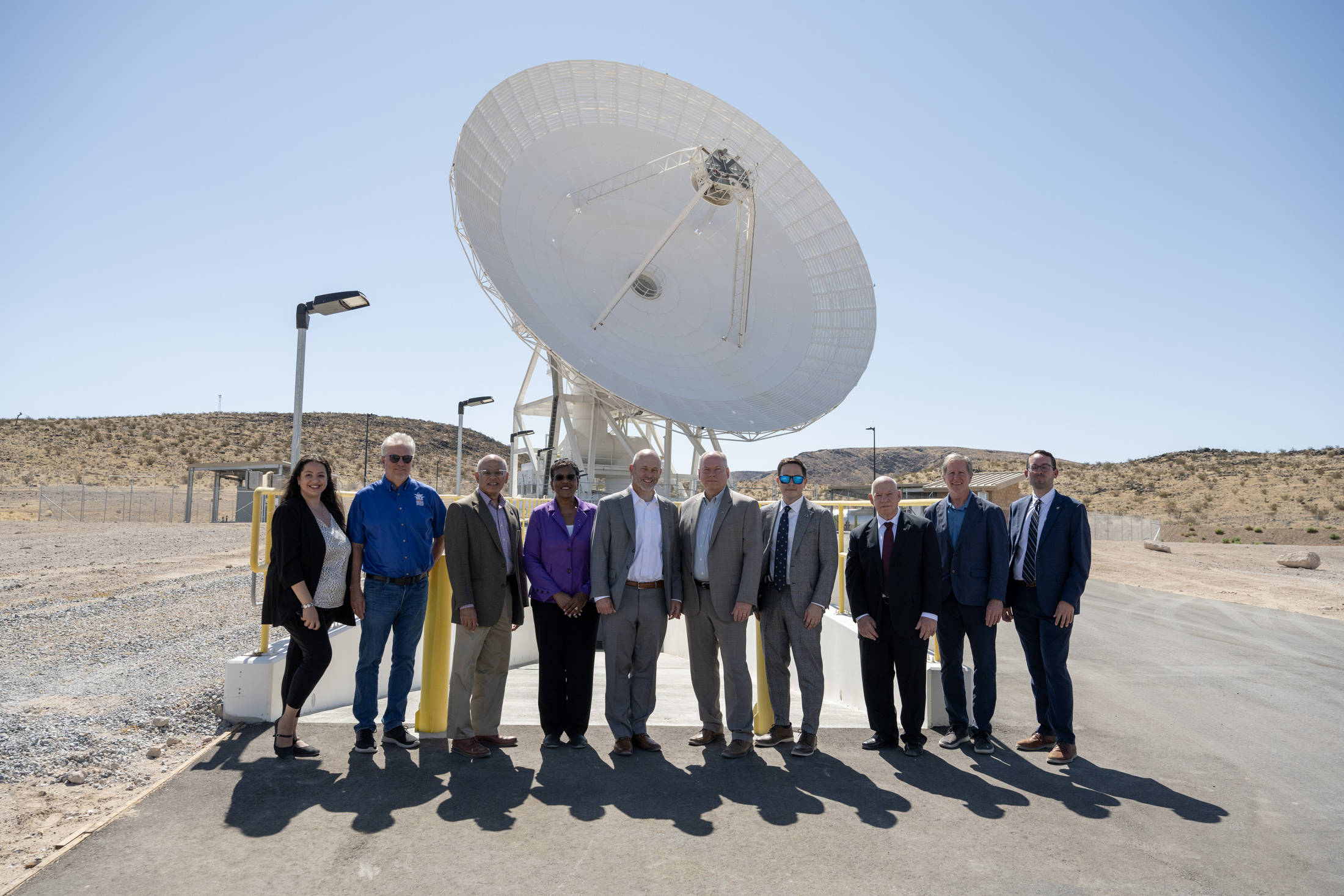 Ten people in professional attire pose together outside under a clear blue sky, with a massive white satellite dish standing directly behind them.