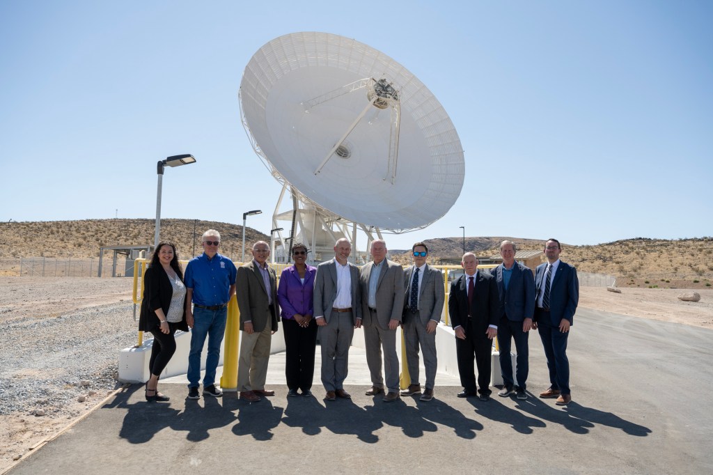 Ten people in professional attire pose together outside under a clear blue sky, with a massive white satellite dish standing directly behind them.