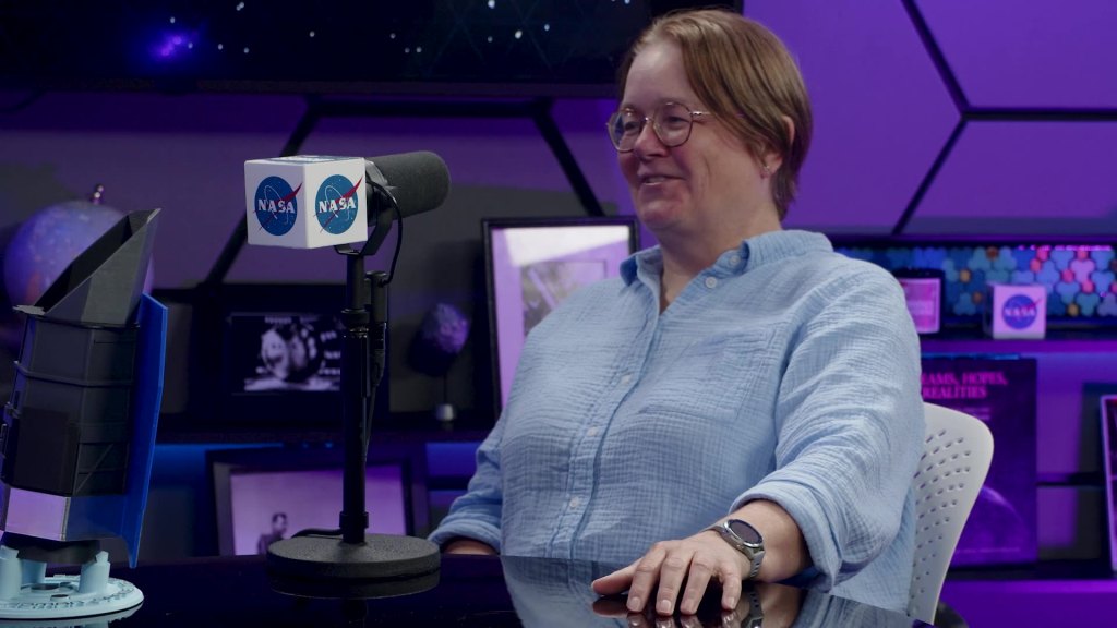 A woman wearing a blue shirt talks into a microphone during a podcast recording. She is sitting at a table with a deep purple background behind her.