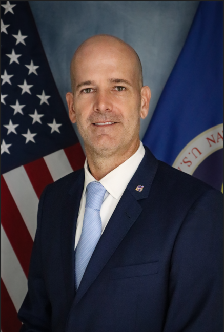 person in a dark blue suit and light blue tie stands in front of an American flag and a blue flag with a circular emblem. The individual has a shaved head, light complexion, and a neutral expression, posing against a studio-style backdrop.