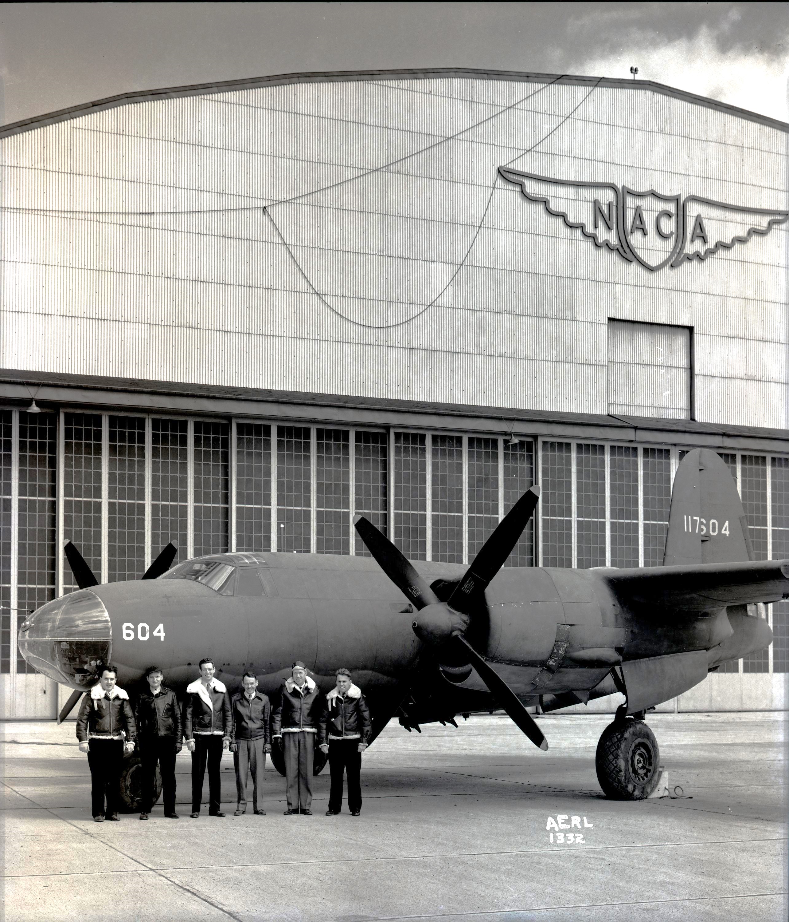 Six men in leather flight jackets stand in front of an airplane that is painted a dull black with the number “604” on the side of the nose. The airplane sits in front of a large hangar with the letters “NACA” on a decorative shield with wings located at the top of the building.