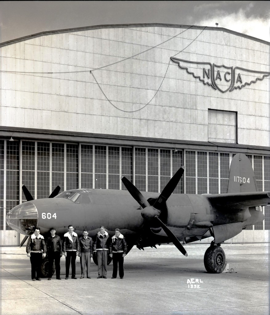 Six men in leather flight jackets stand in front of an airplane that is painted a dull black with the number “604” on the side of the nose. The airplane sits in front of a large hangar with the letters “NACA” on a decorative shield with wings located at the top of the building.