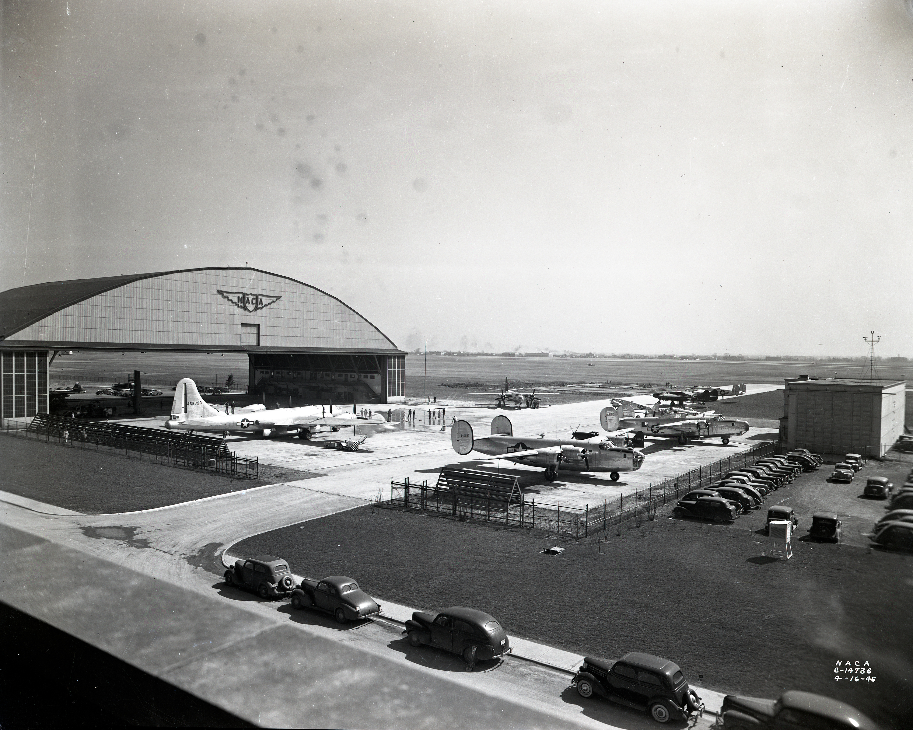 Several airplanes parked outside a large hangar with cars in a nearby parking lot on an airfield.