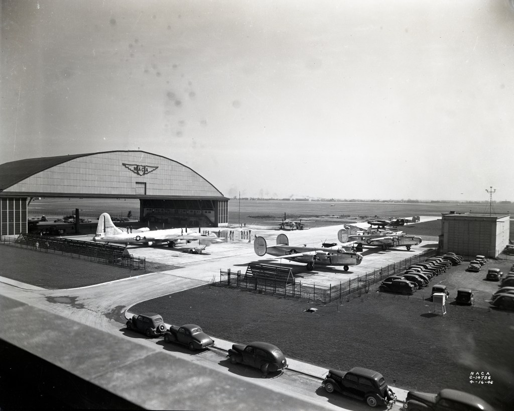 Several airplanes parked outside a large hangar with cars in a nearby parking lot on an airfield.