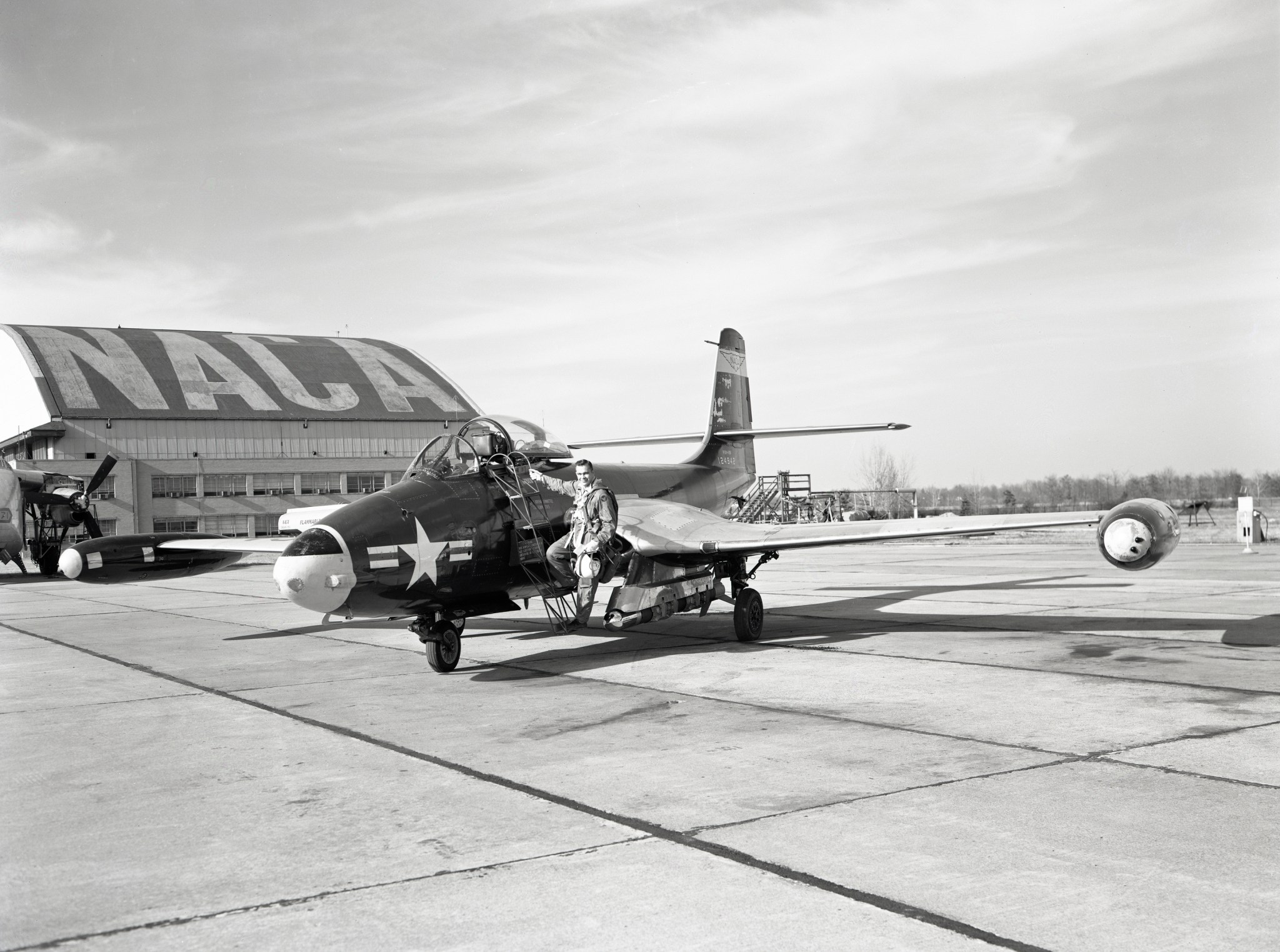 A pilot steps on a metal ladder to get onto a small, one-seater airplane. The NACA hangar is in background