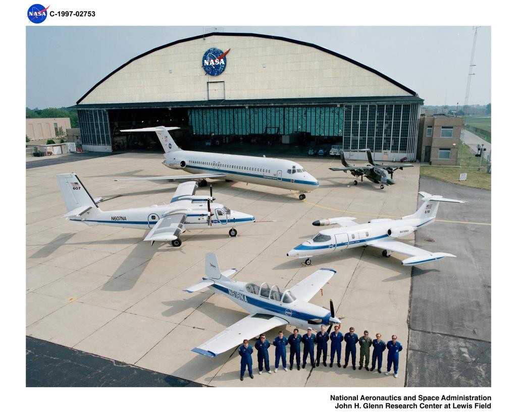A fleet of five aircraft are strategically situated on the tarp of NASA’s Glenn (Lewis) Research Center hanger, which is pictured in the background. Eleven staff members in flight suits with their arms behind their backs stand in the foreground.