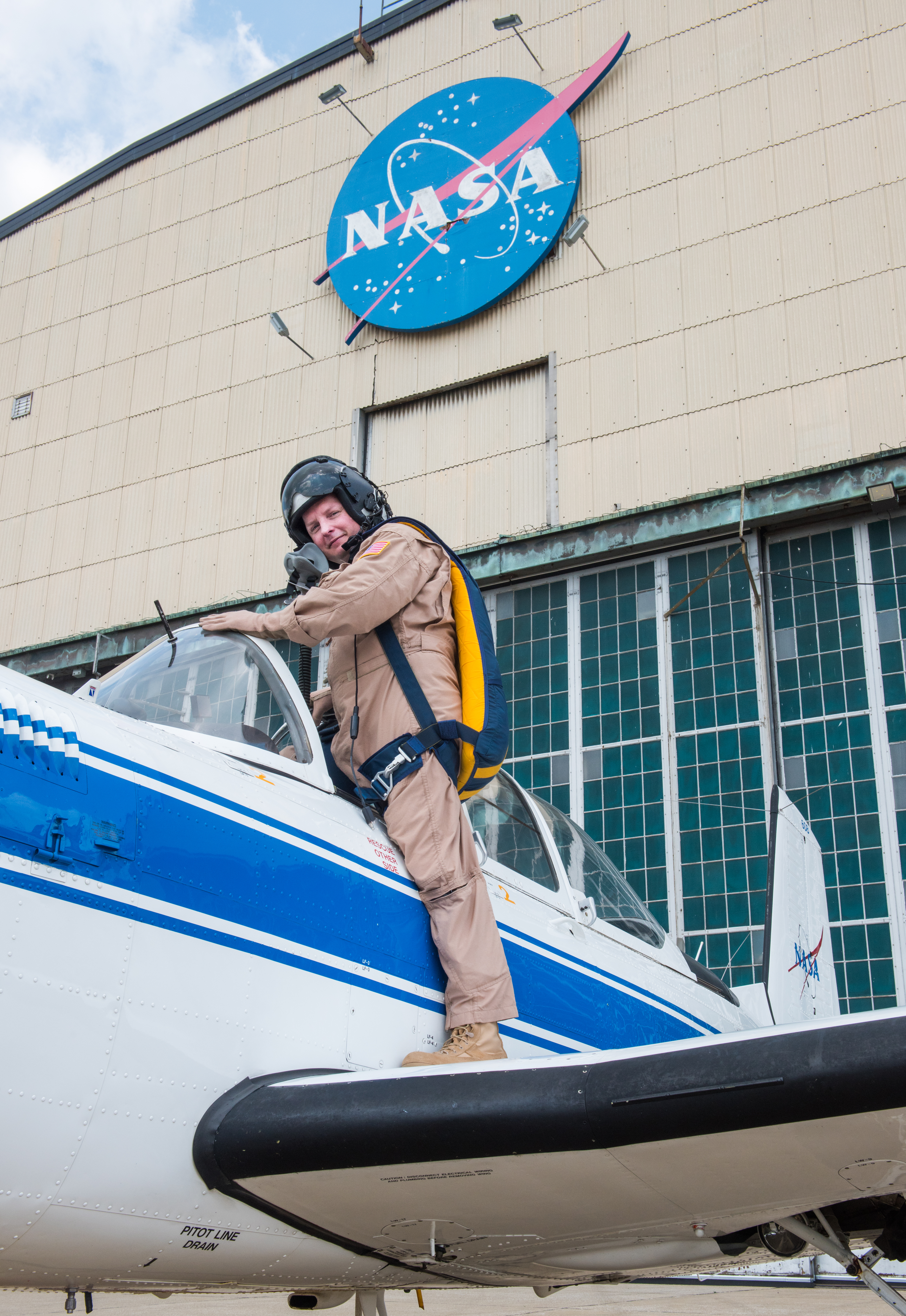 A pilot in a flight suit stands on the wing of a small airplane with one leg and climbs inside the cockpit with the other. The NASA Glenn hangar is in the background.