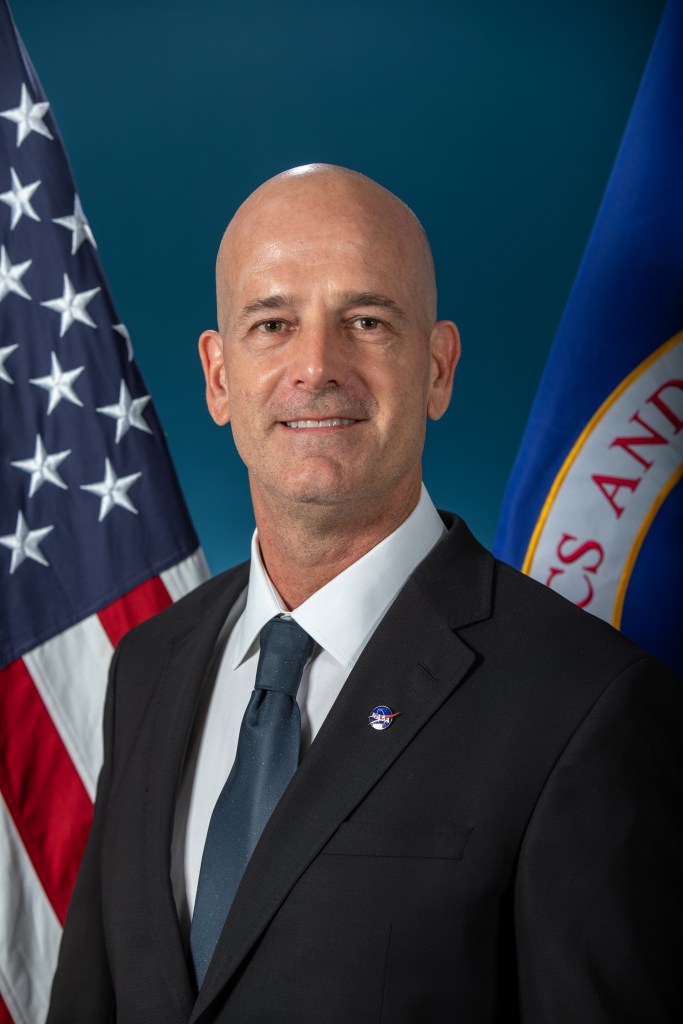 Image shows man posing for photograph wearing a black suit and dark tie with a NASA pin on his suit. In the background, an American flag and NASA flag.