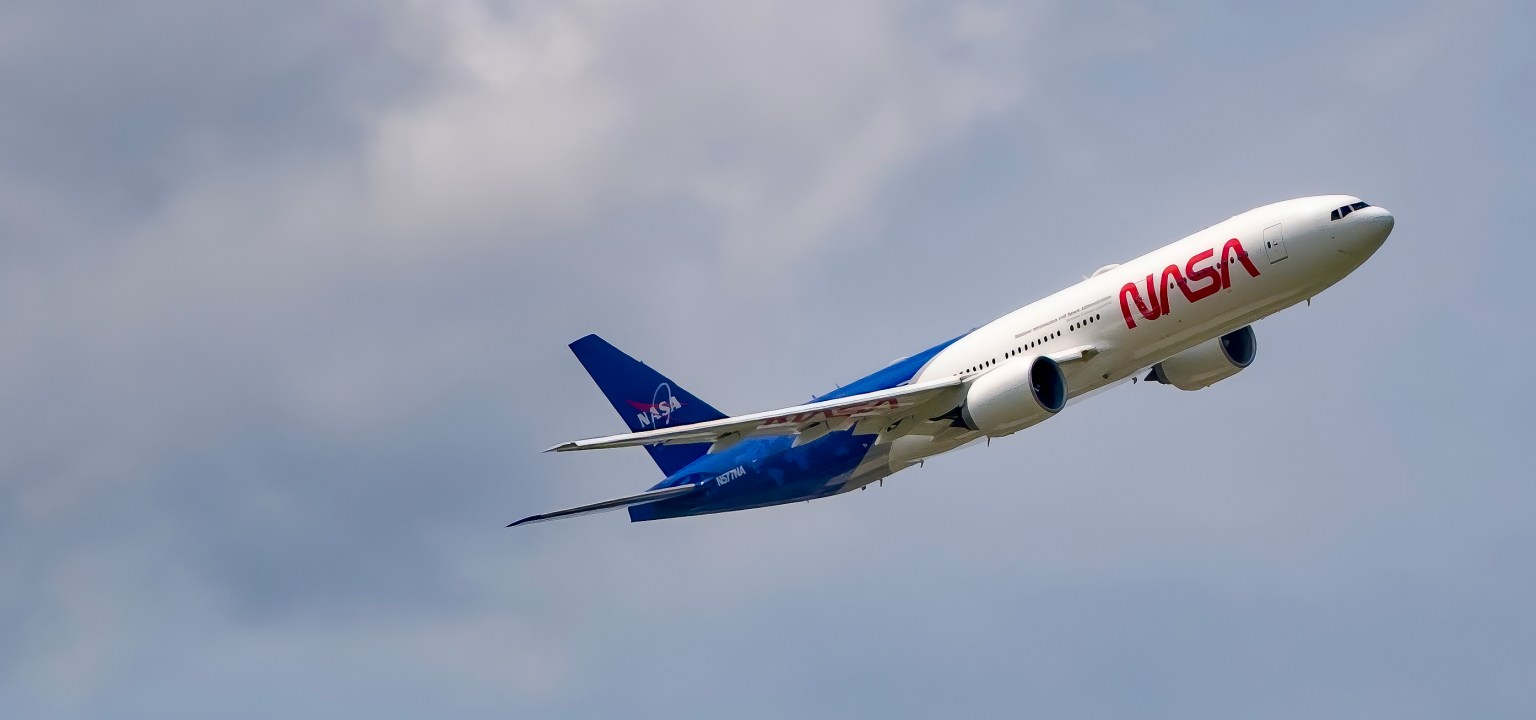 A large blue and white airliner with two engines mounted to its wings sports NASA logos as it flies overhead.
