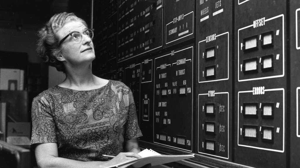 An archival, black and white photo of Nancy Grace Roman. She is holding papers and looking up at a board full of information