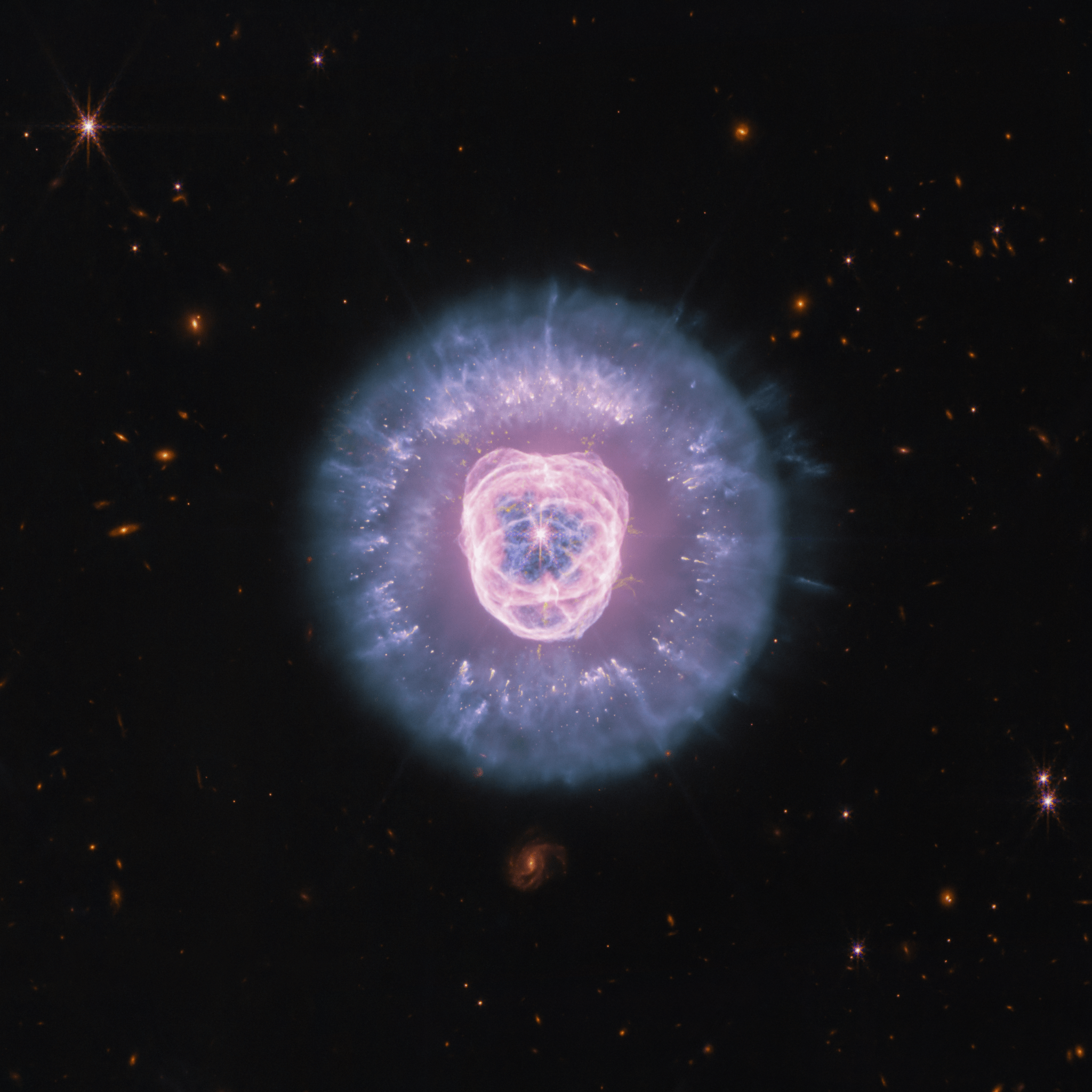 JWST’s Near Infrared Camera and Mid Infrared Instrument combined image of planetary nebula NGC 2392, the Lion Nebula, against the black background of space. The nebula is at center, circular in shape, and looks like a male lion’s head. In the center is a small, pinkish-white circle with eight-point diffraction spikes, a white dwarf star. Surrounding the star are purple-pink cavernous bubbles and shell-like rings. The bubbles and shells collectively form an oval with two small, wide arcs near the top, reminiscent of a lion’s face and its ears. What appears to be extended outward from the lion’s face is a thick ring of purple-blue material. The width of the ring is consistent throughout and resembles a mane. The area closest to the lion’s face looks cloudy, while the edges of the mane look like there are clumps of dust with comet-like tails. In the background are distant galaxies and stars. Some galaxies are yellow and orange points of light and others have spiral structures. Some stars have diffraction spikes.