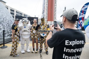 Three Pittsburgh Steelers fans, dressed in black and yellow clothes with the Steelers' logo on it, pose with an astronaut in a spacesuit. The reflective visor is closed. In the foreground, a man takes the group's picture with his phone.