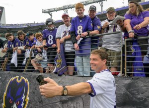 Reid Wiseman holds up a black phone to take a selfie with Baltimore Ravens fans at a football game. The fans are dressed in Ravens jerseys and shirts in purple and white. The fans are in the stands above, while Wiseman is on the field just below.