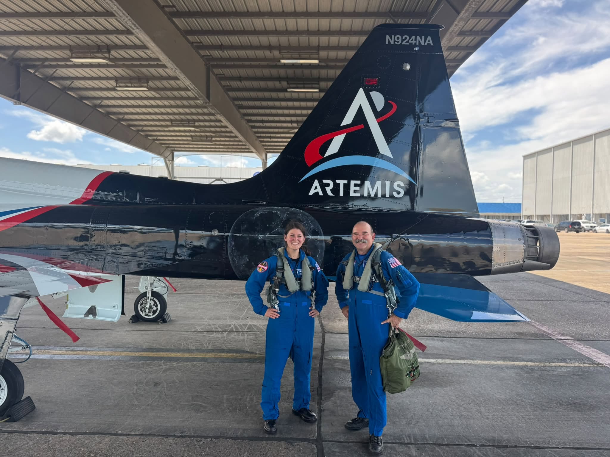 A woman and a man in blue flight suits stand in front of a T-38 jet with NASA's Artemis program logo on the tail.