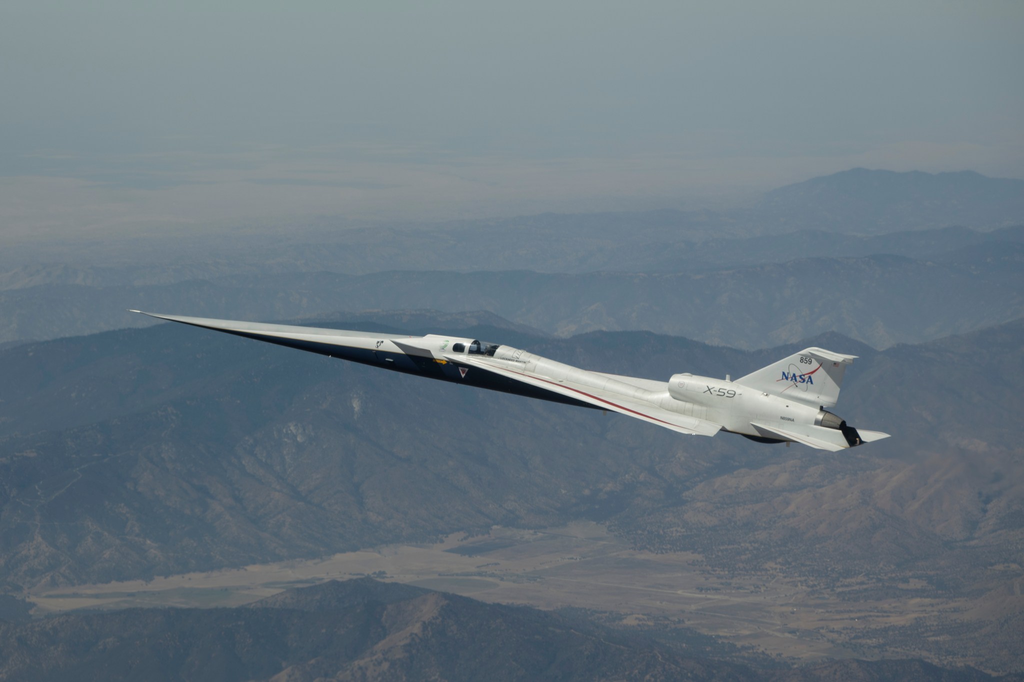 NASA’s white and blue X-59 quiet supersonic experimental aircraft flies above mountainous terrain in the Mojave Desert. The aircraft is shown from the side with its long nose extending toward the left.