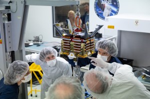 A group of five people wearing cleanroom suits, hairnets, and masks watch a small gold and silver box suspended by a small crane in a laboratory. In the background, slightly out of focus, two more people watch through a window. Next to the window is a large decal of the COSI mission patch.