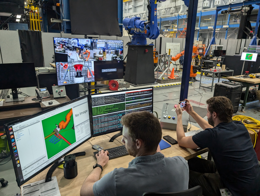 Two men sit at a wooden table with two large computer monitors displaying data and imagery from a robotic arm test. They sit in a large, open facility with additional computer monitors and robotic equipment in the background.