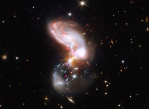 In this system, two galaxies are merging at a furious rate. Near the top of the image is a spiral galaxy shape with thick arms in fiery oranges, whites, and reds. At the bottom of the image is a faint hazy bowl shape marbled with grainy white ribbons and hot pink specks. Where the two shapes collide, in the center of the frame, the galaxy appears utterly chaotic, like a firework.
