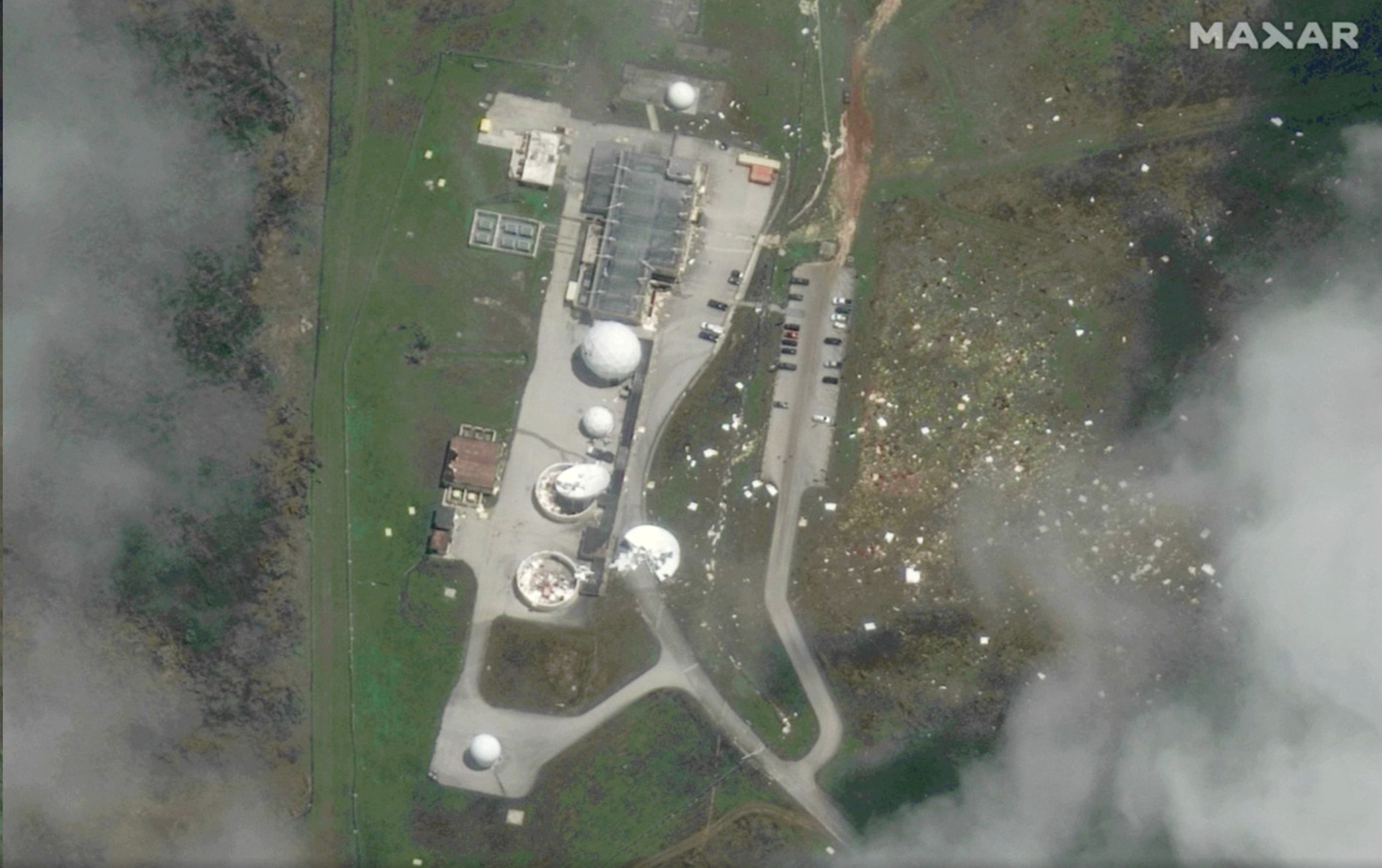 Aerial view of the same communications facility after severe storm damage, with several radomes torn open or damaged and debris scattered across the surrounding grounds.