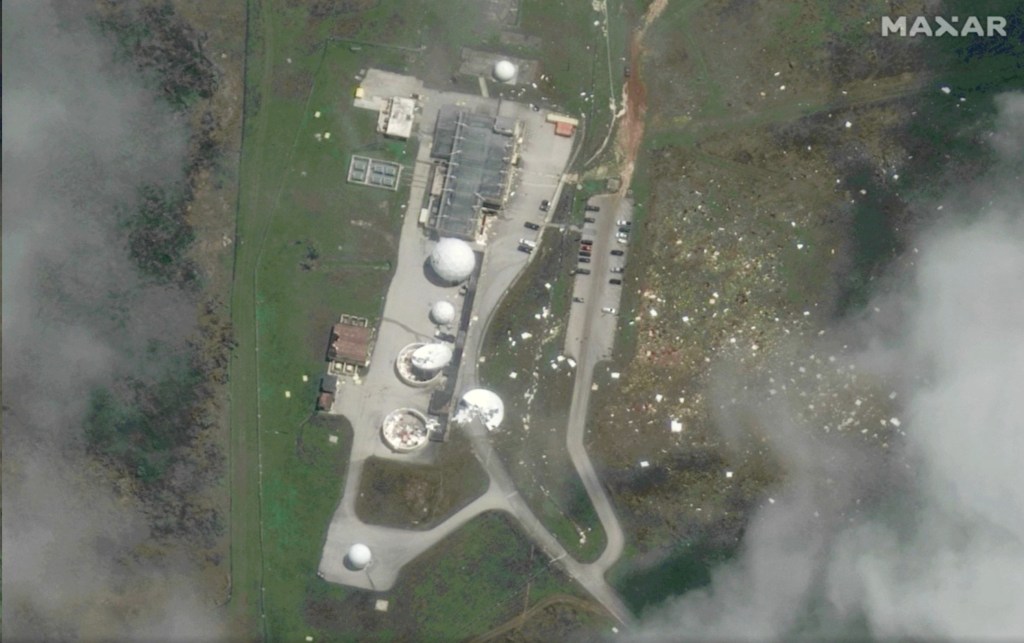 Aerial view of the same communications facility after severe storm damage, with several radomes torn open or damaged and debris scattered across the surrounding grounds.