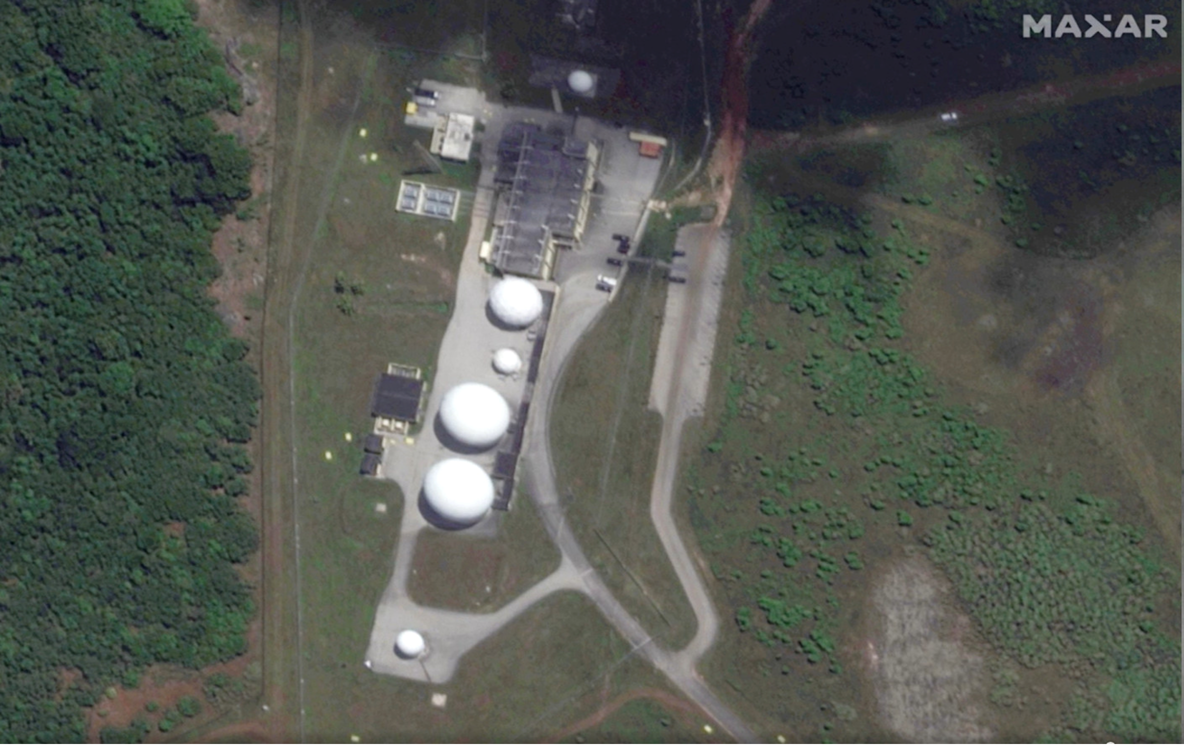 Aerial view of a communications facility surrounded by dense vegetation, showing several white radomes, support buildings, and an access road.