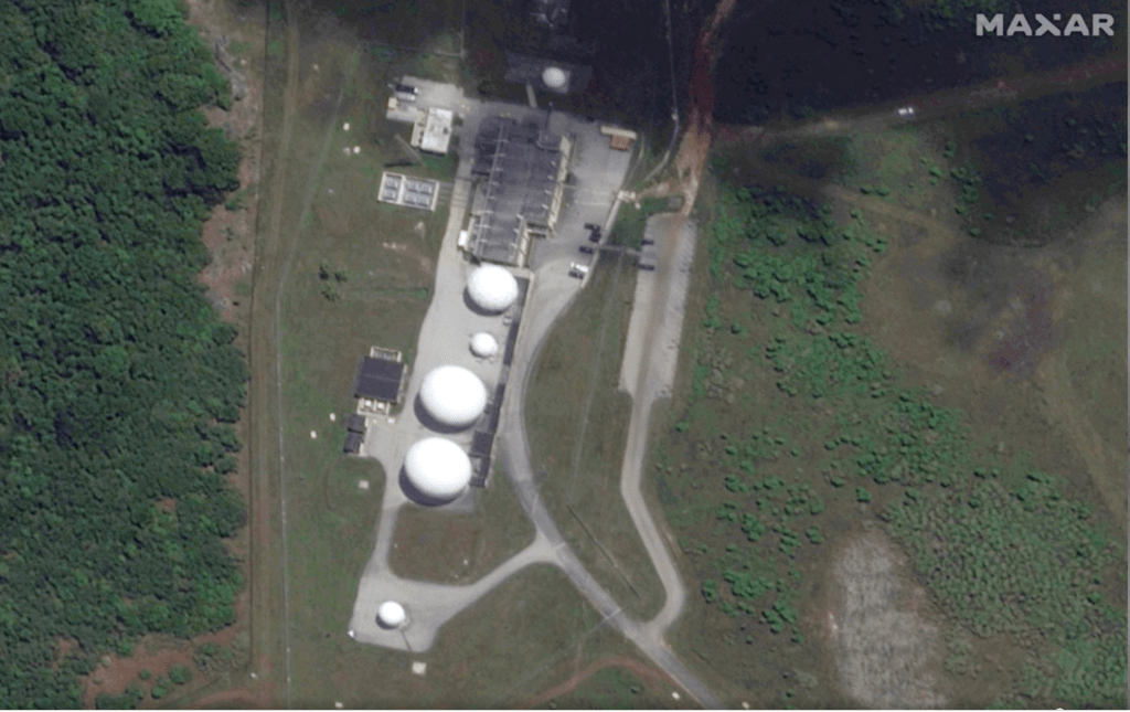 Aerial view of a communications facility surrounded by dense vegetation, showing several white radomes, support buildings, and an access road.