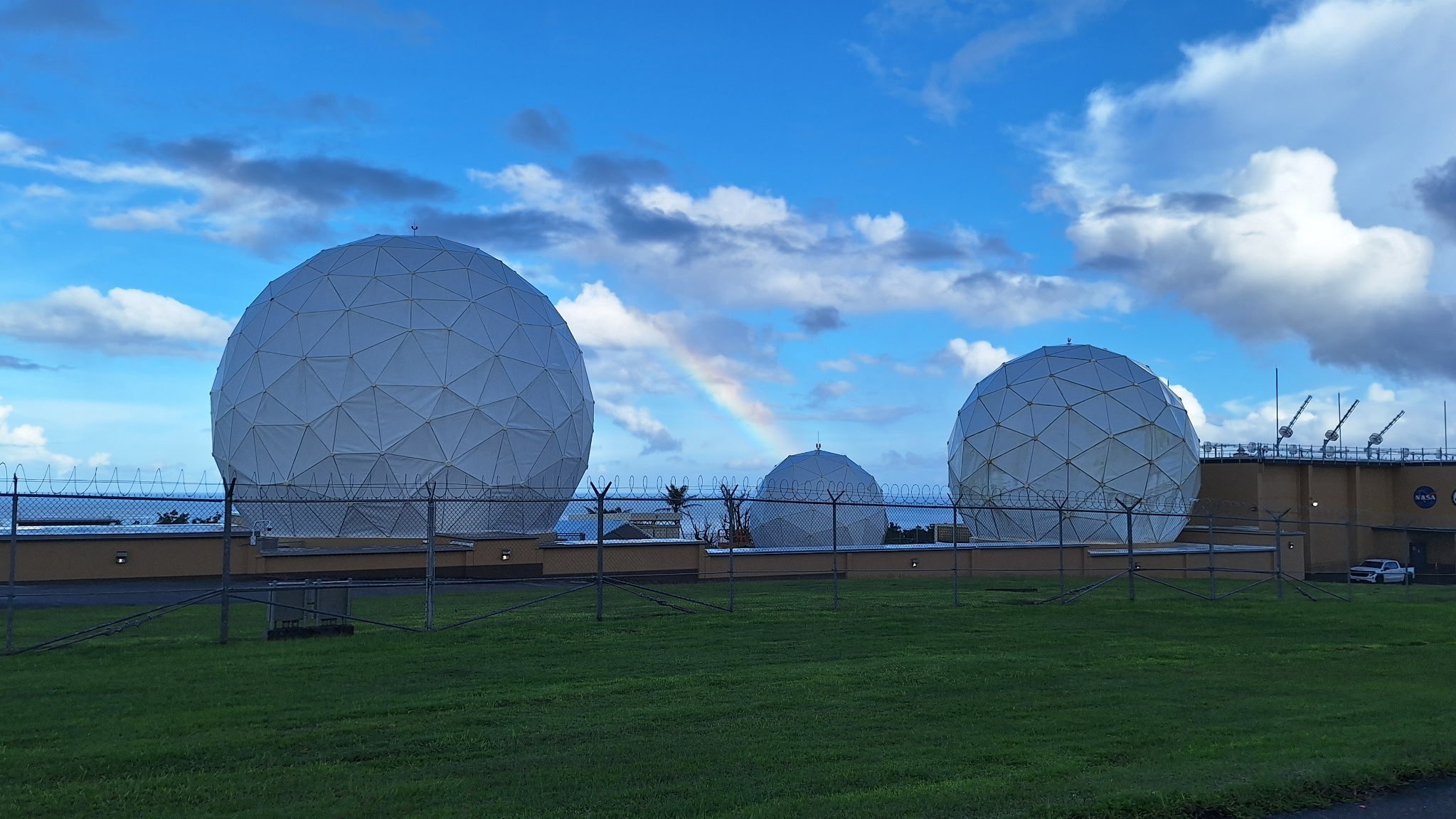 Three large white spherical radio antennas at the repaired Guam Remote Station stand behind a security fence on a grassy field. A rainbow arcs through the partly cloudy blue sky behind the antennas.