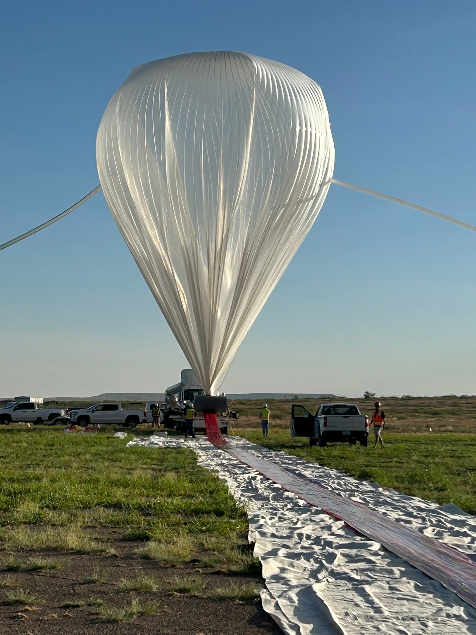 A scientific balloon partially inflated before flight. The balloon is clear plastic in the shape of an upside down teardrop. The rest of the ballon trails behind it on the ground.
