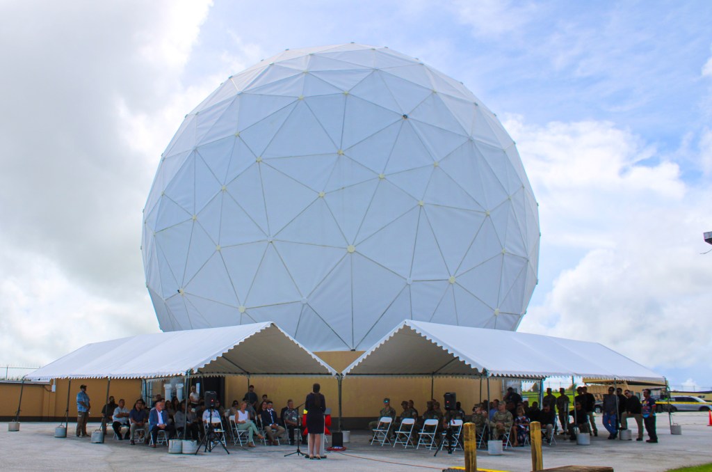 Employees and guests gather beneath white event tents outside a large white geodesic radome during an outdoor ceremony under partly cloudy skies. A woman in a dark suit addresses the audience.