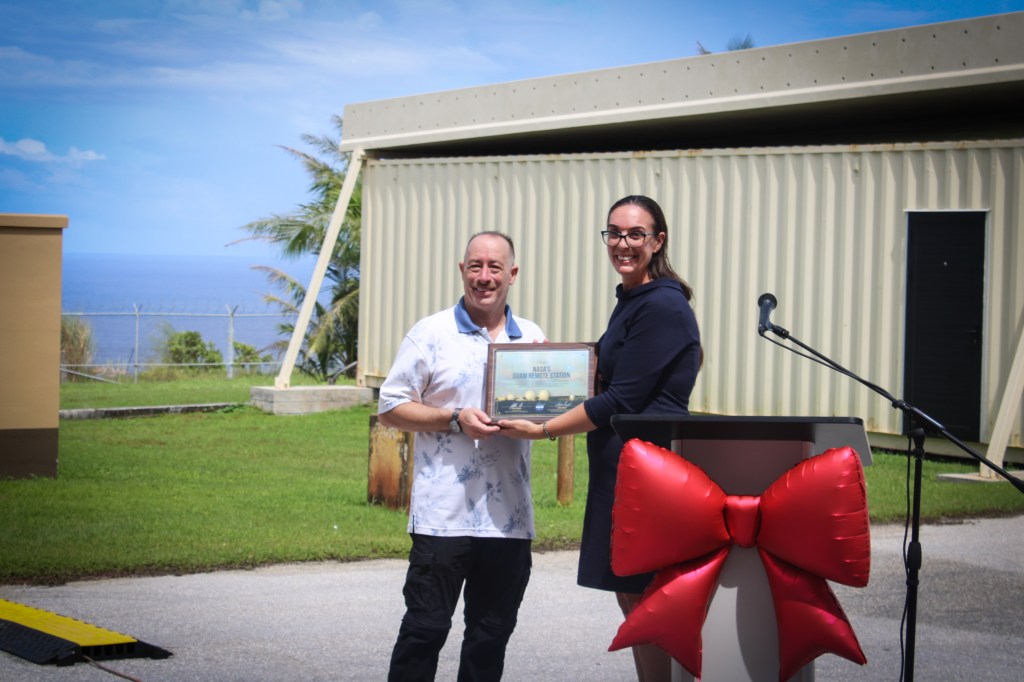 Two people smile while holding a framed award during an outdoor ceremony; a podium decorated with a large red bow stands beside them, with facility buildings and the ocean in the background.