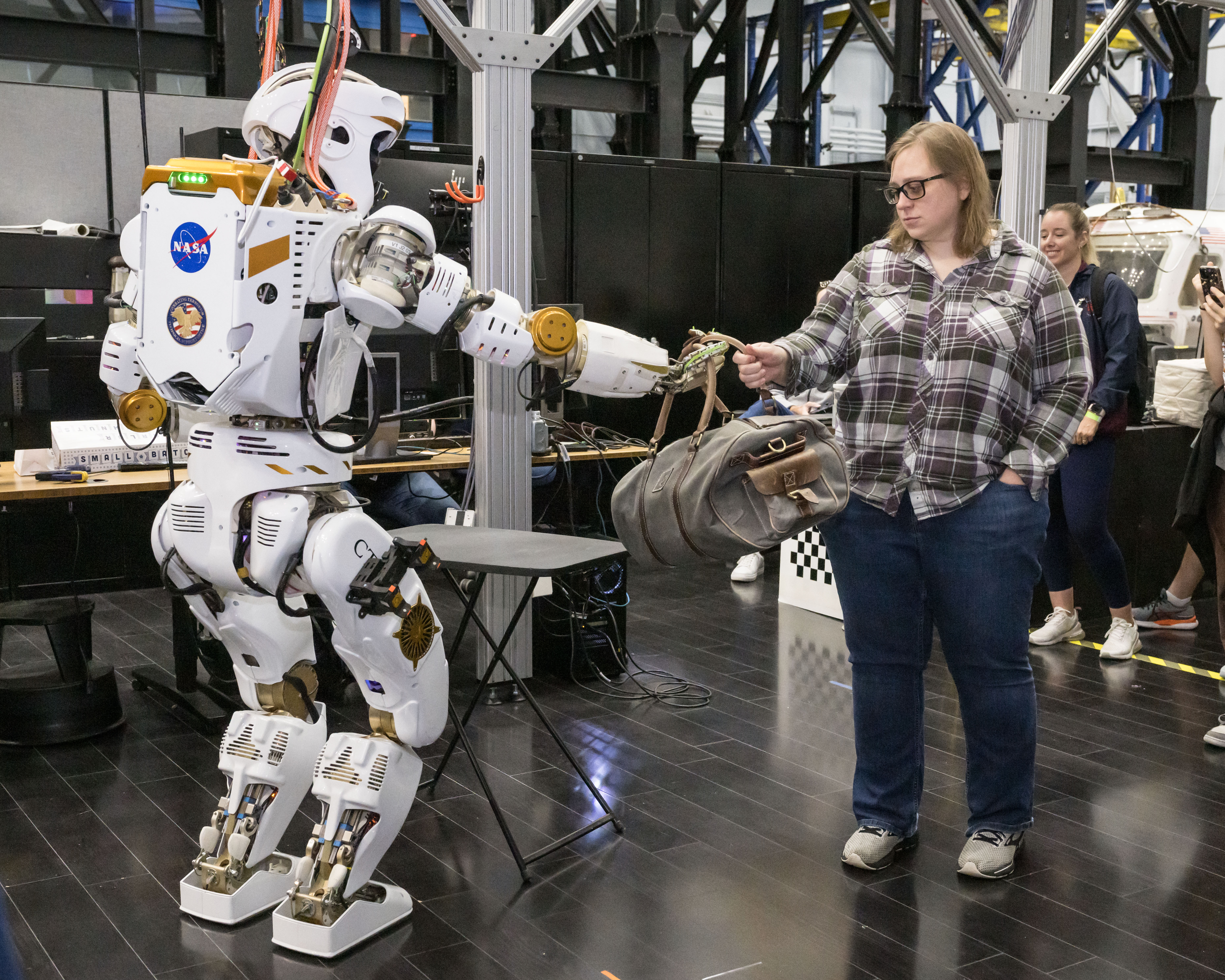 A humanoid robot hands a duffel bag to a woman wearing casual clothes.