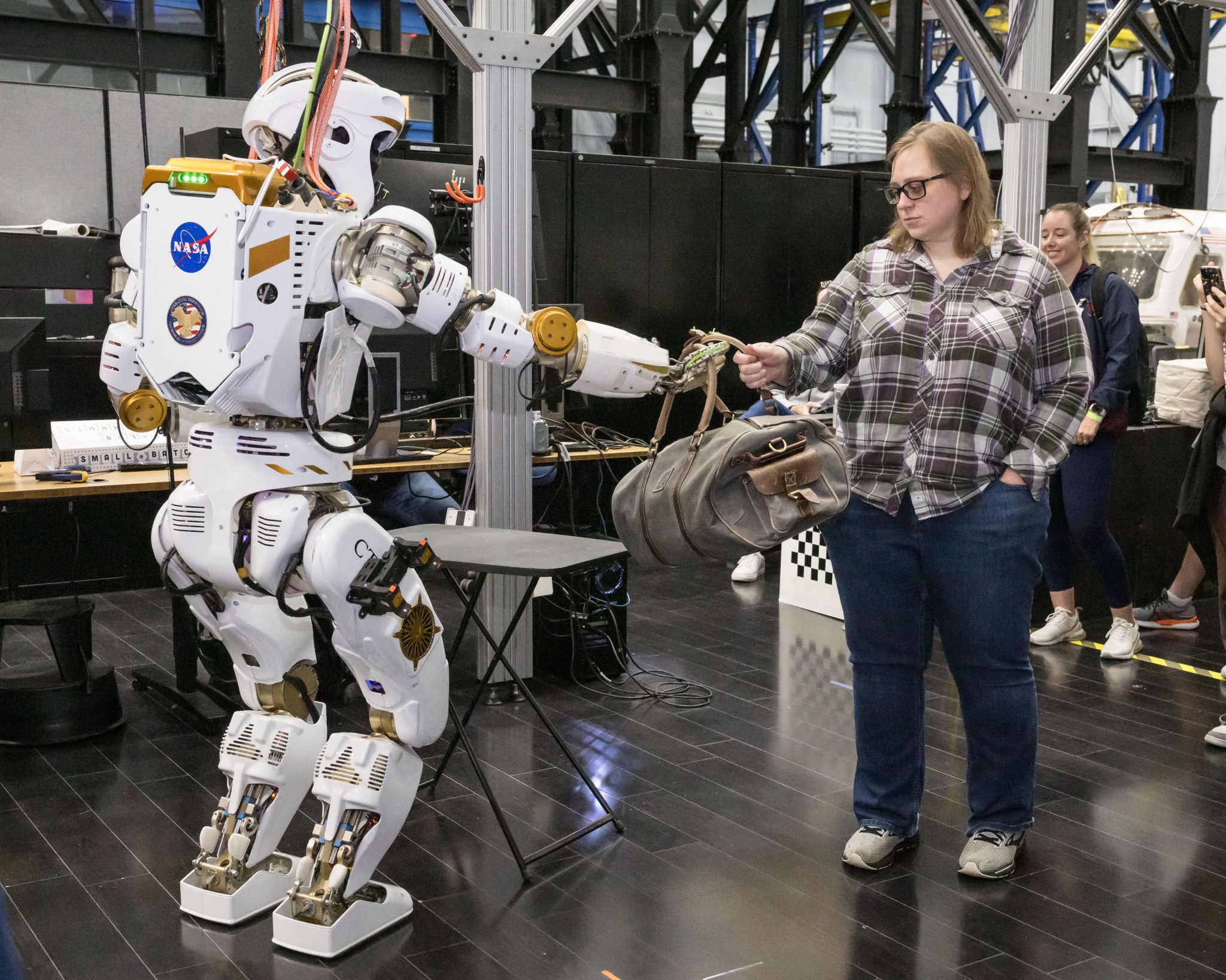 A humanoid robot hands a duffel bag to a woman wearing casual clothes.