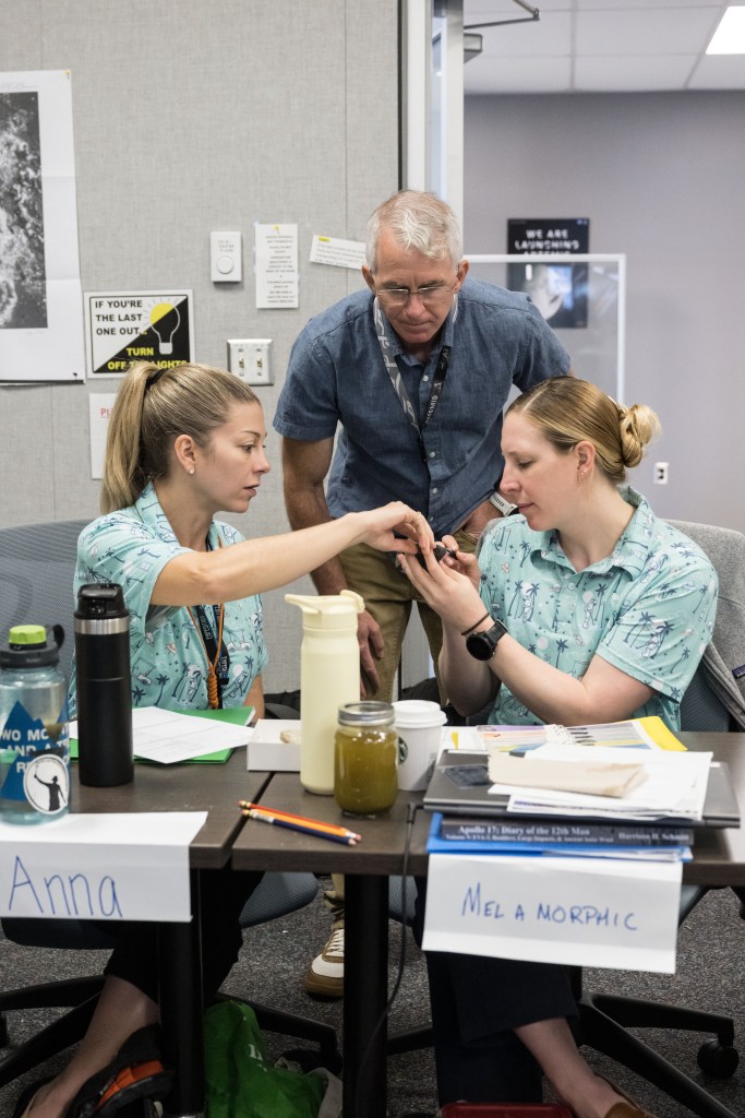 Two women wearing matching button-down shirts study a rock sample with an instructor looking over them in the background.