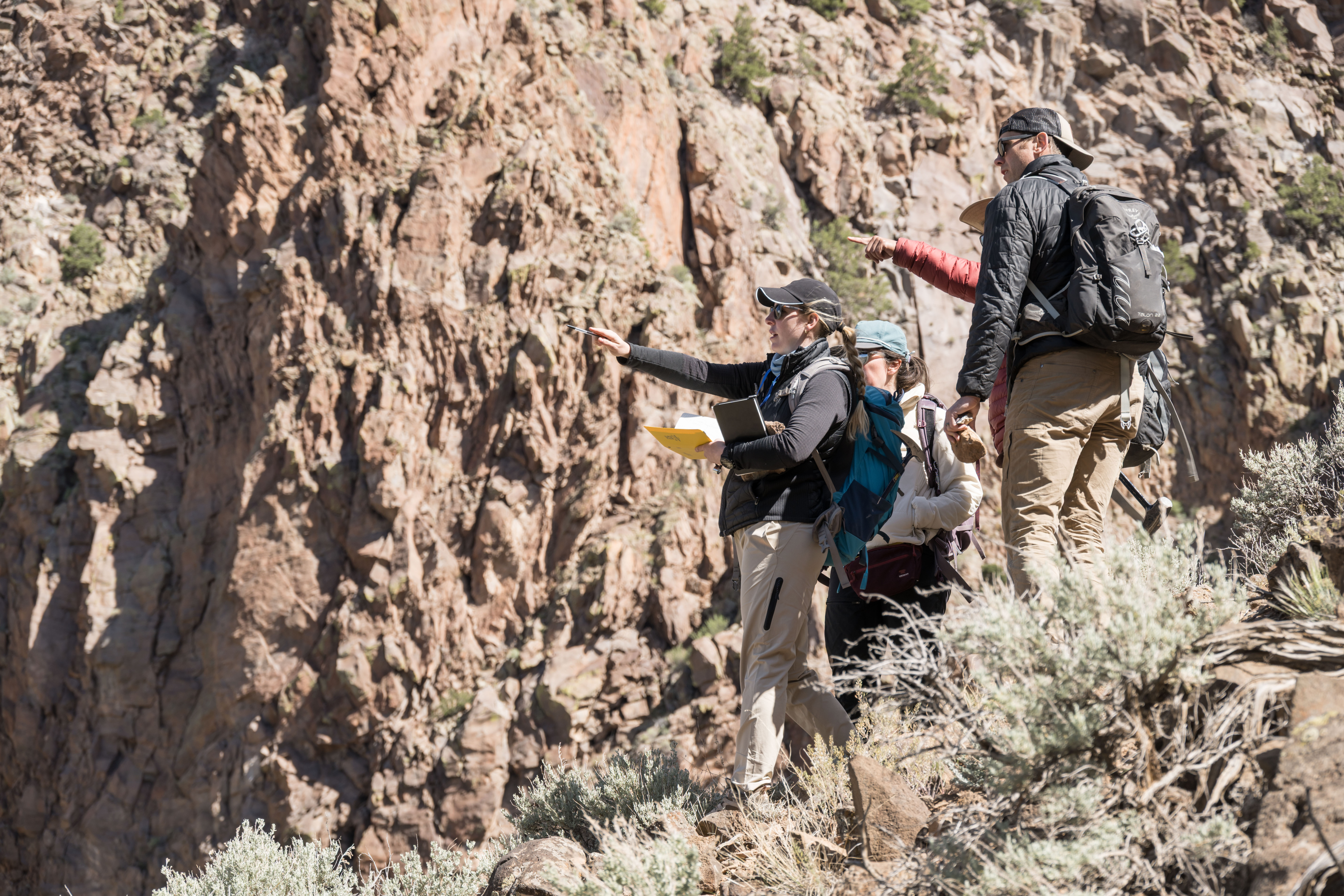 A group of four men and women dressed in hiking attire pause on a rocky hillside to point at something off-camera.