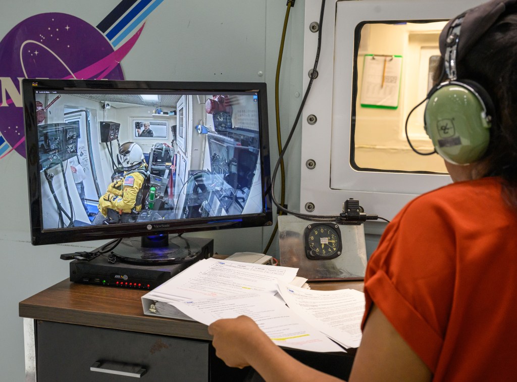 A woman wearing earmuffs watches a computer monitor showing a man in a yellow flight suit sitting inside an altitude chamber.
