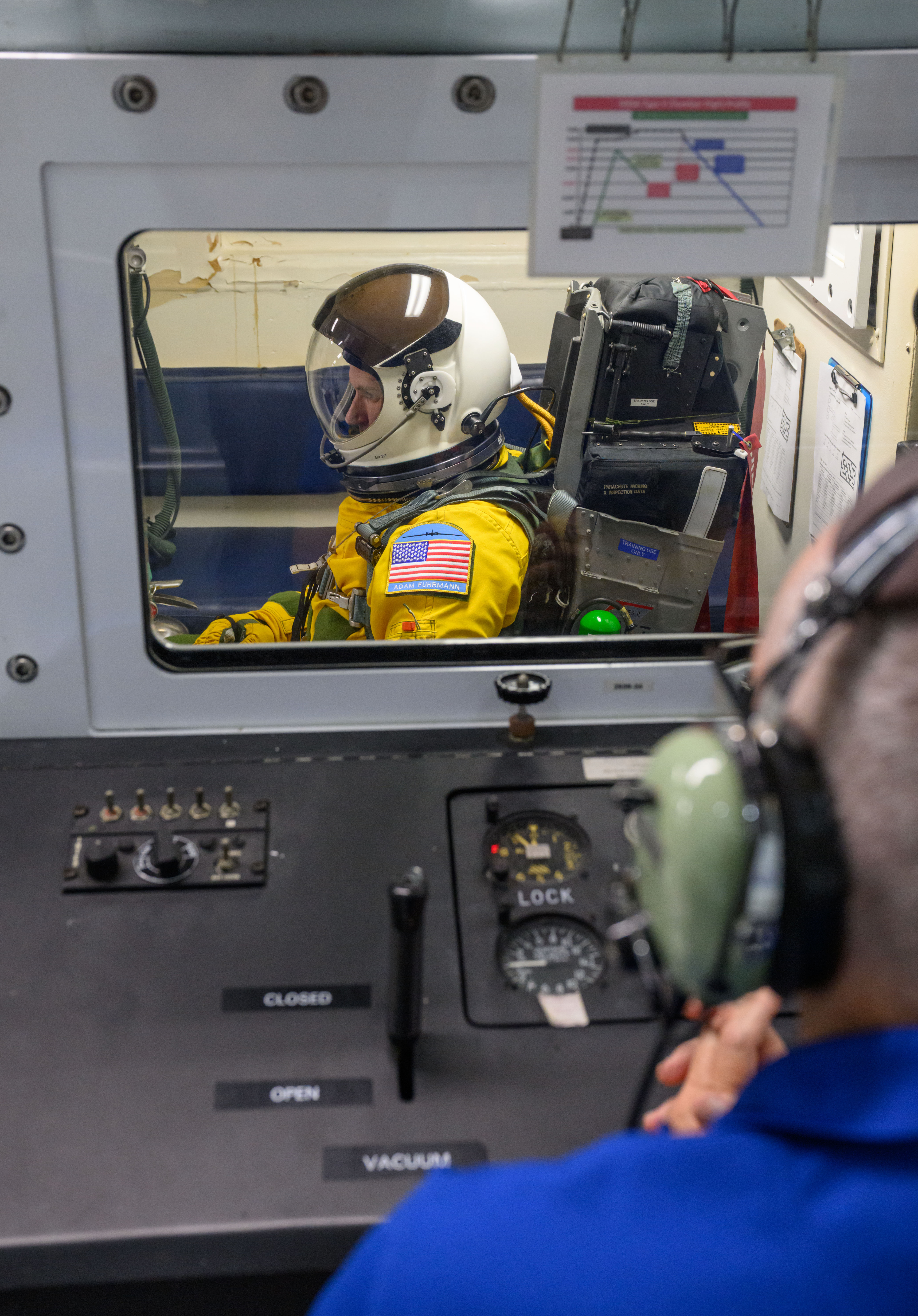 A man wearing a yellow flight suit and a white helmet sits inside an altitude chamber.