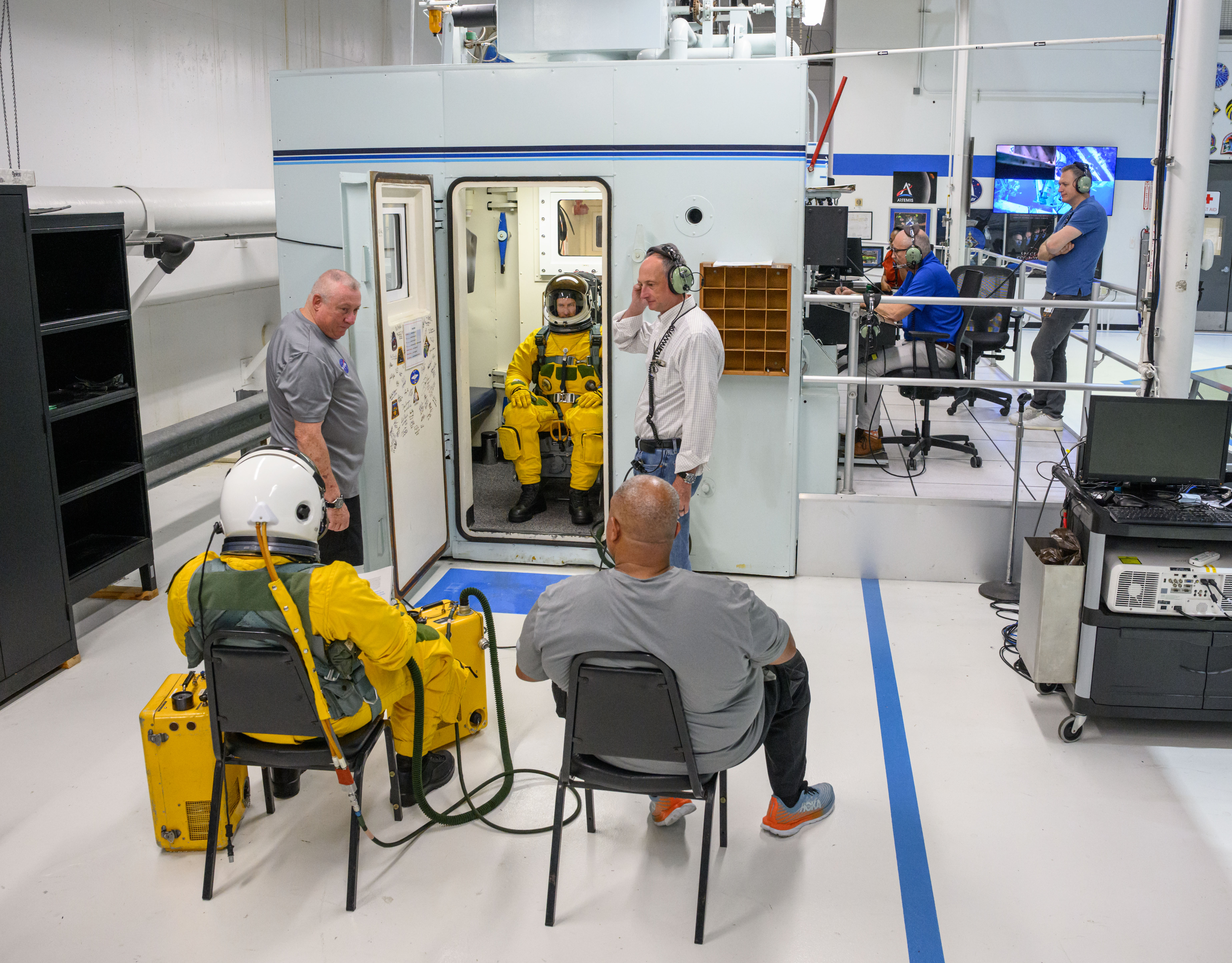 A person wearing a yellow flight suit and white helmet sits inside an altitude chamber with the door propped open, while four technicians, one instructor, and another individual wearing a yellow flight suit observes the proceedings.