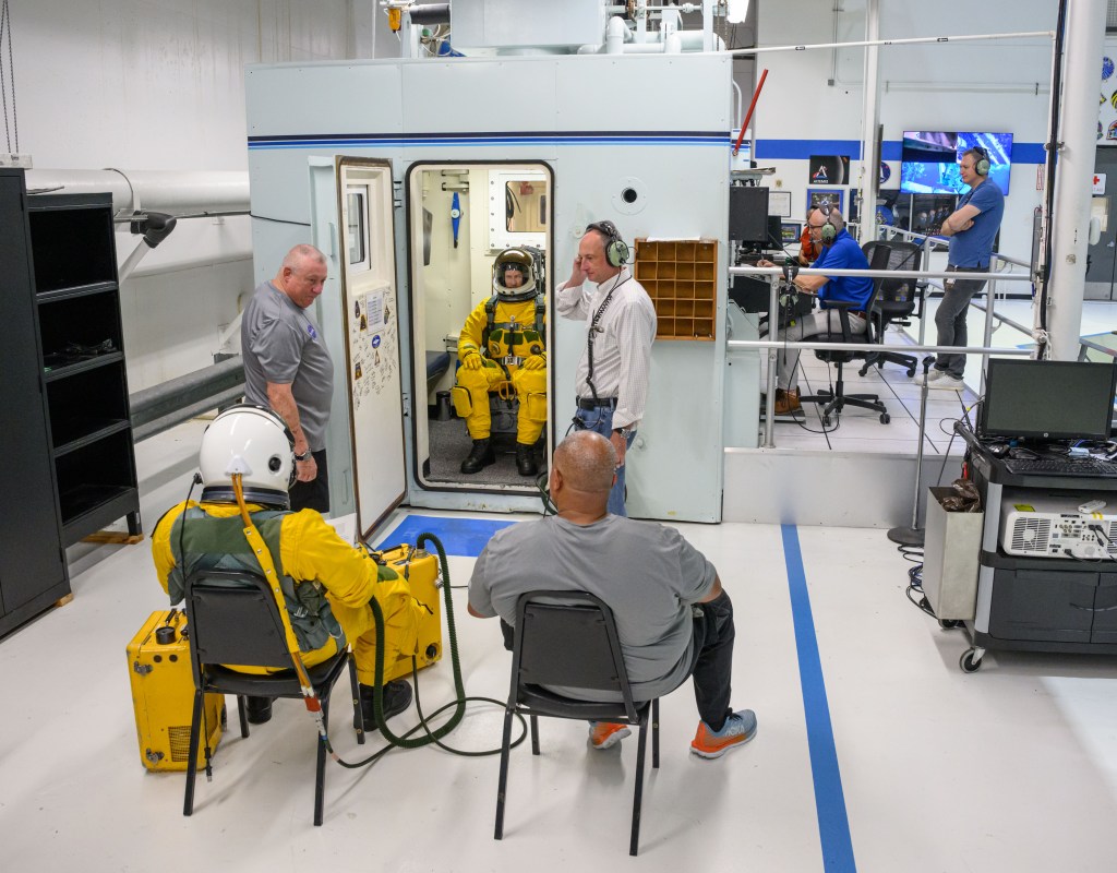A person wearing a yellow flight suit and white helmet sits inside an altitude chamber with the door propped open, while four technicians, one instructor, and another individual wearing a yellow flight suit observes the proceedings.