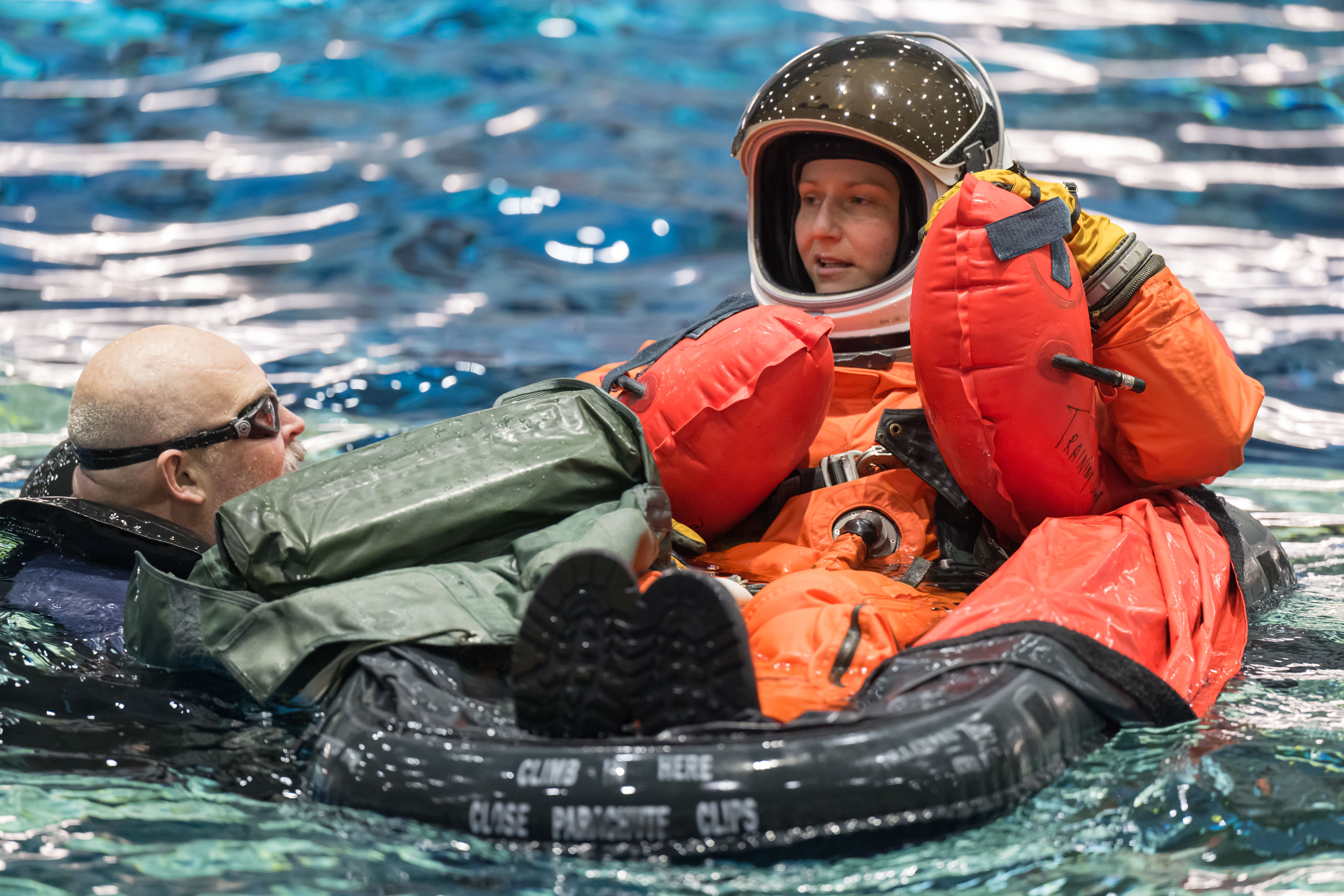 A woman in an orange flight suit and white helmet floats inside a small raft while speaking to a diver at the Neutral Buoyancy Laboratory.