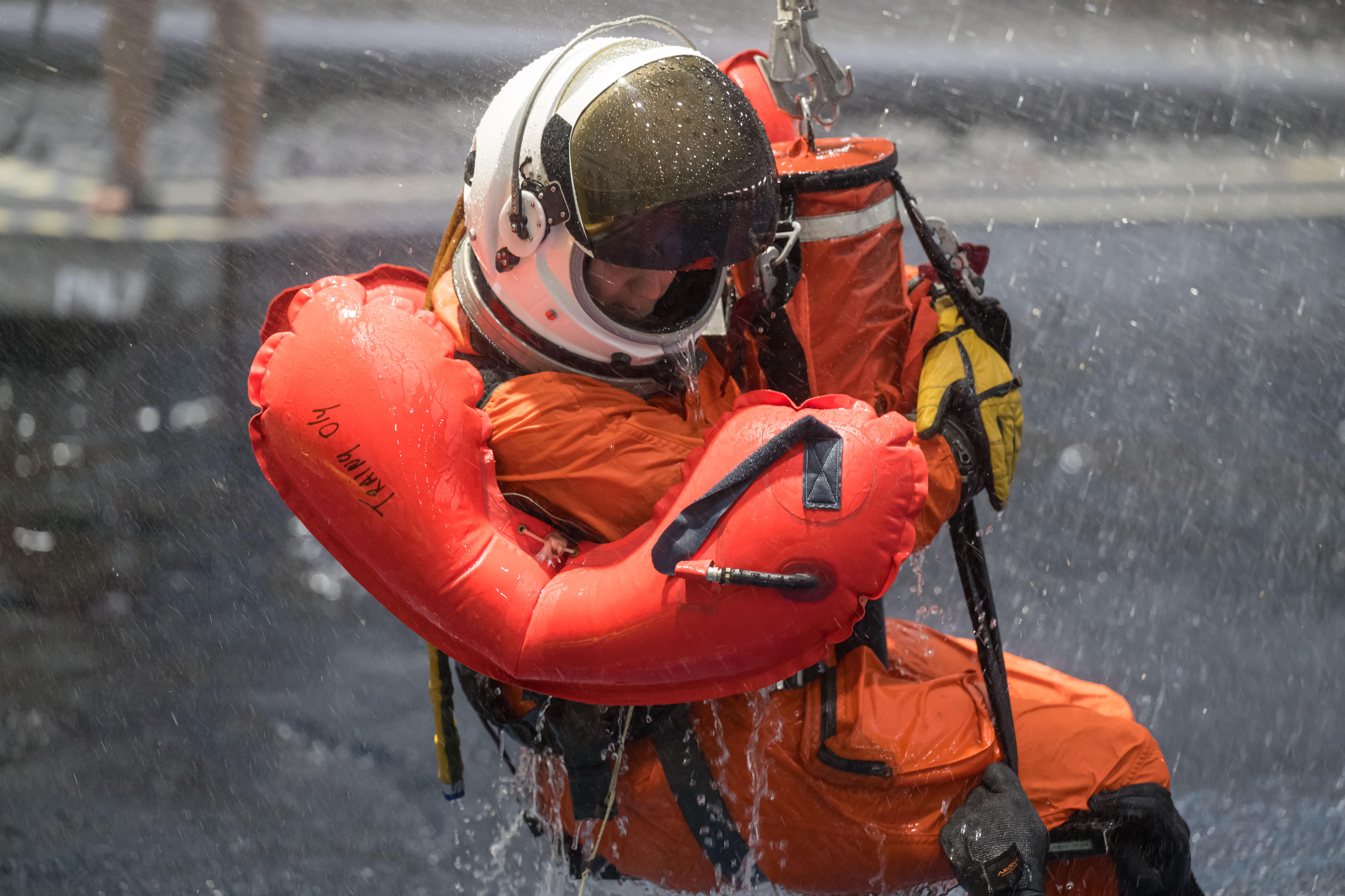 A person wearing an orange flight suit and white helmet is lifted from the Neutral Buoyancy Laboratory pool with a flotation device secured around their waist.